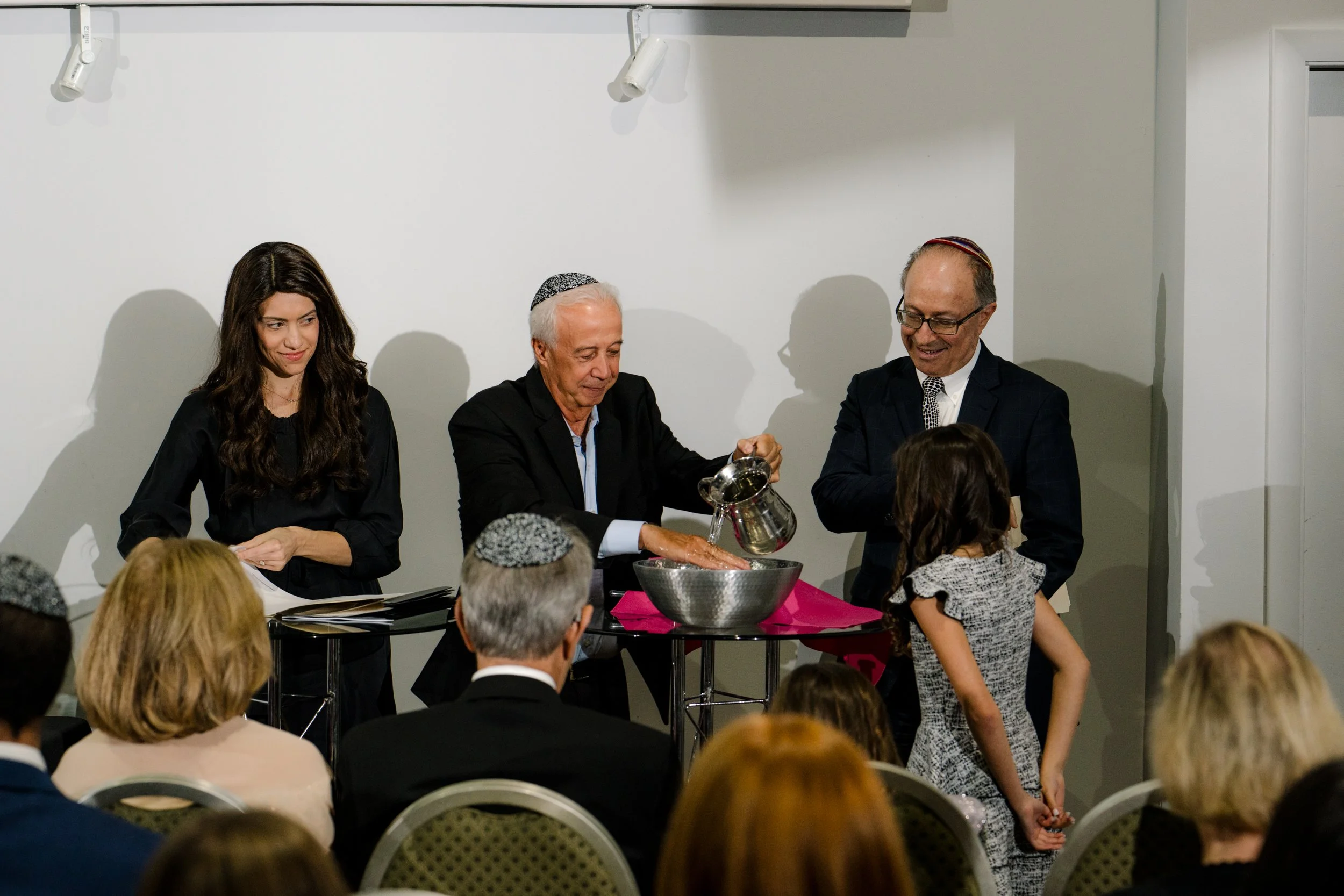 A Jewish ceremony where a man pours water from a pitcher into a bowl, surrounded by people wearing yarmulkes in a formal setting.