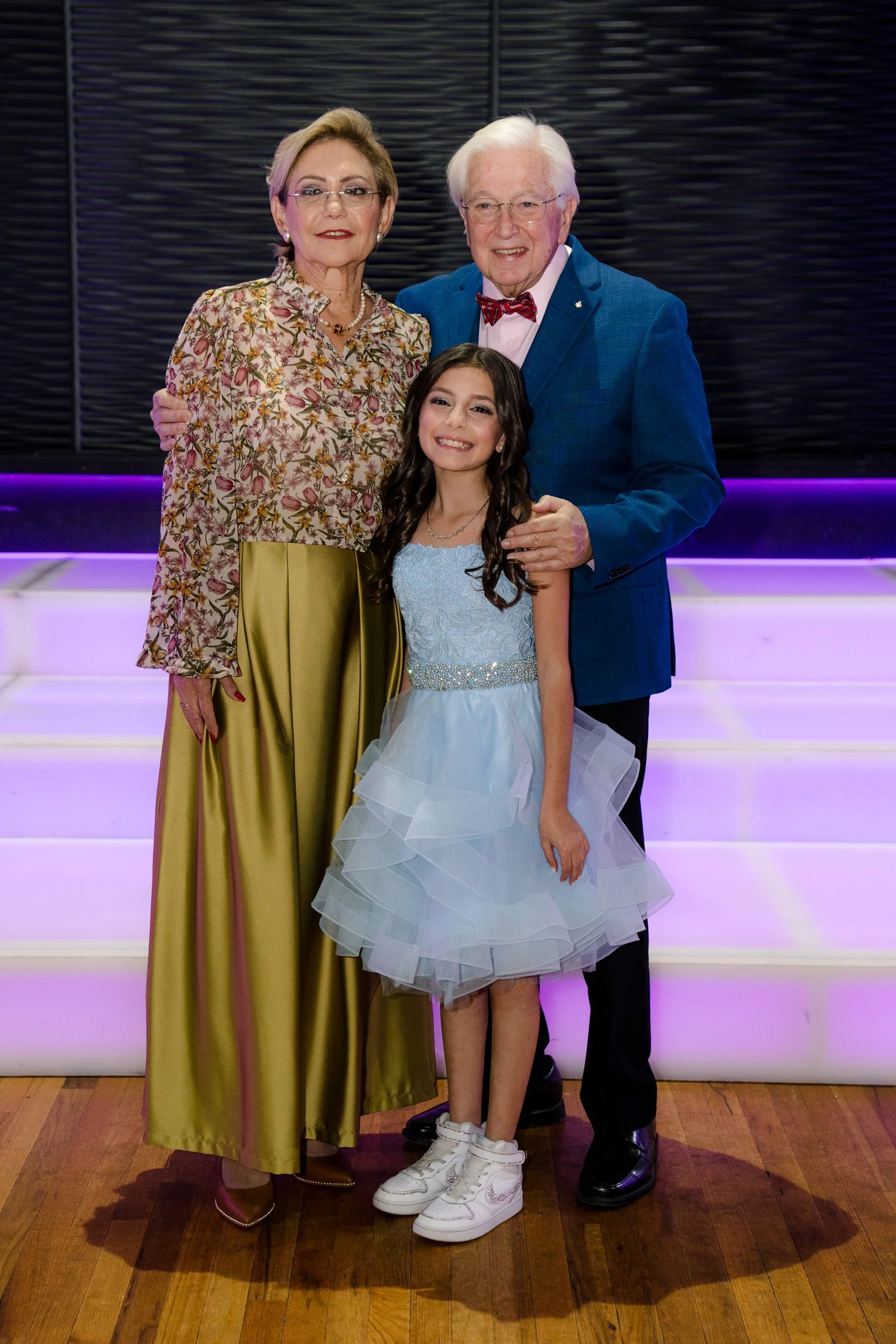 Three people posing together: an elderly man in a blue suit and red bow tie, a woman in a floral blouse and gold skirt, and a young girl in a light blue dress and white sneakers.