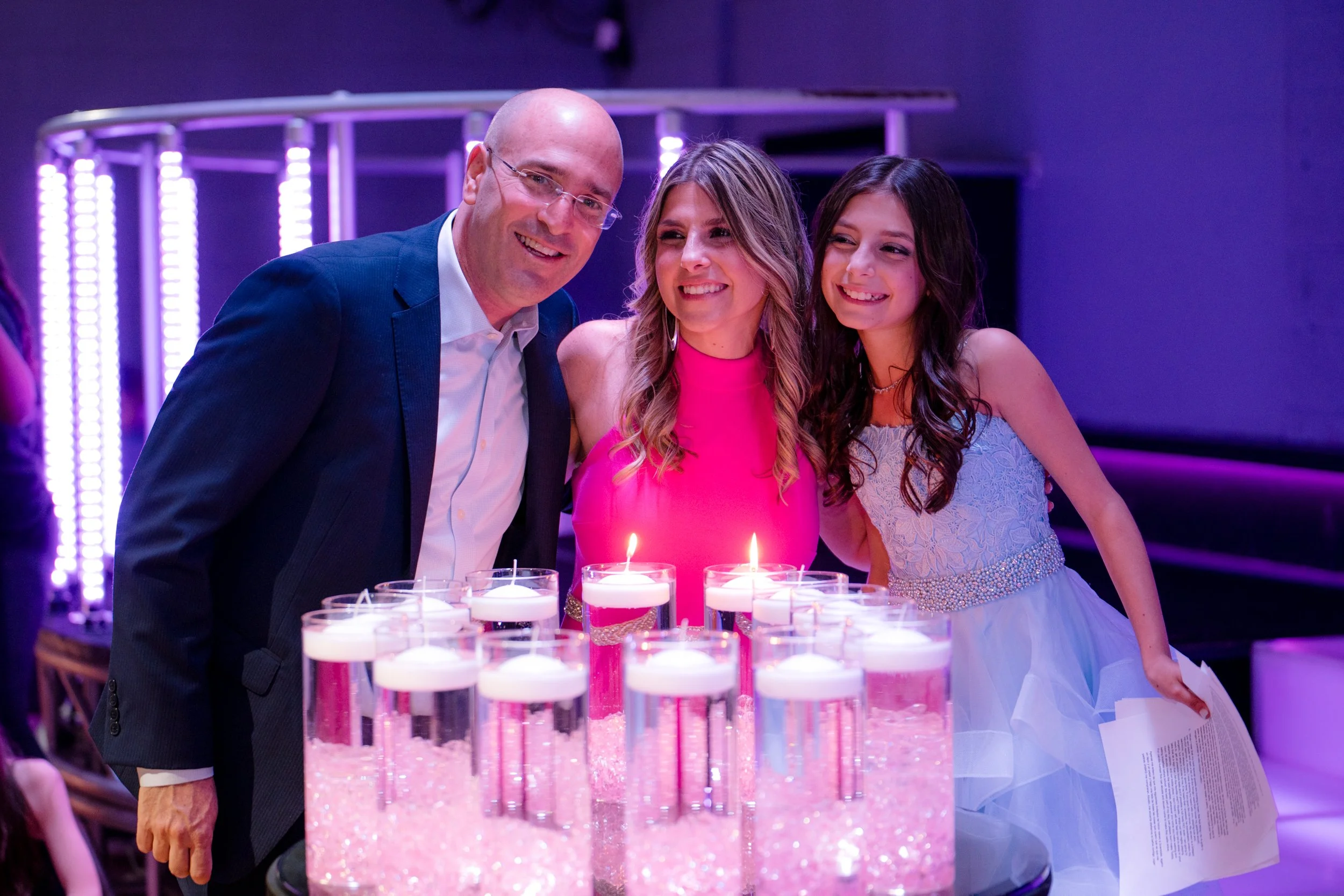 Three people smiling at an event with floating candles in glass vases. The background is lit with purple lights, creating a festive atmosphere.