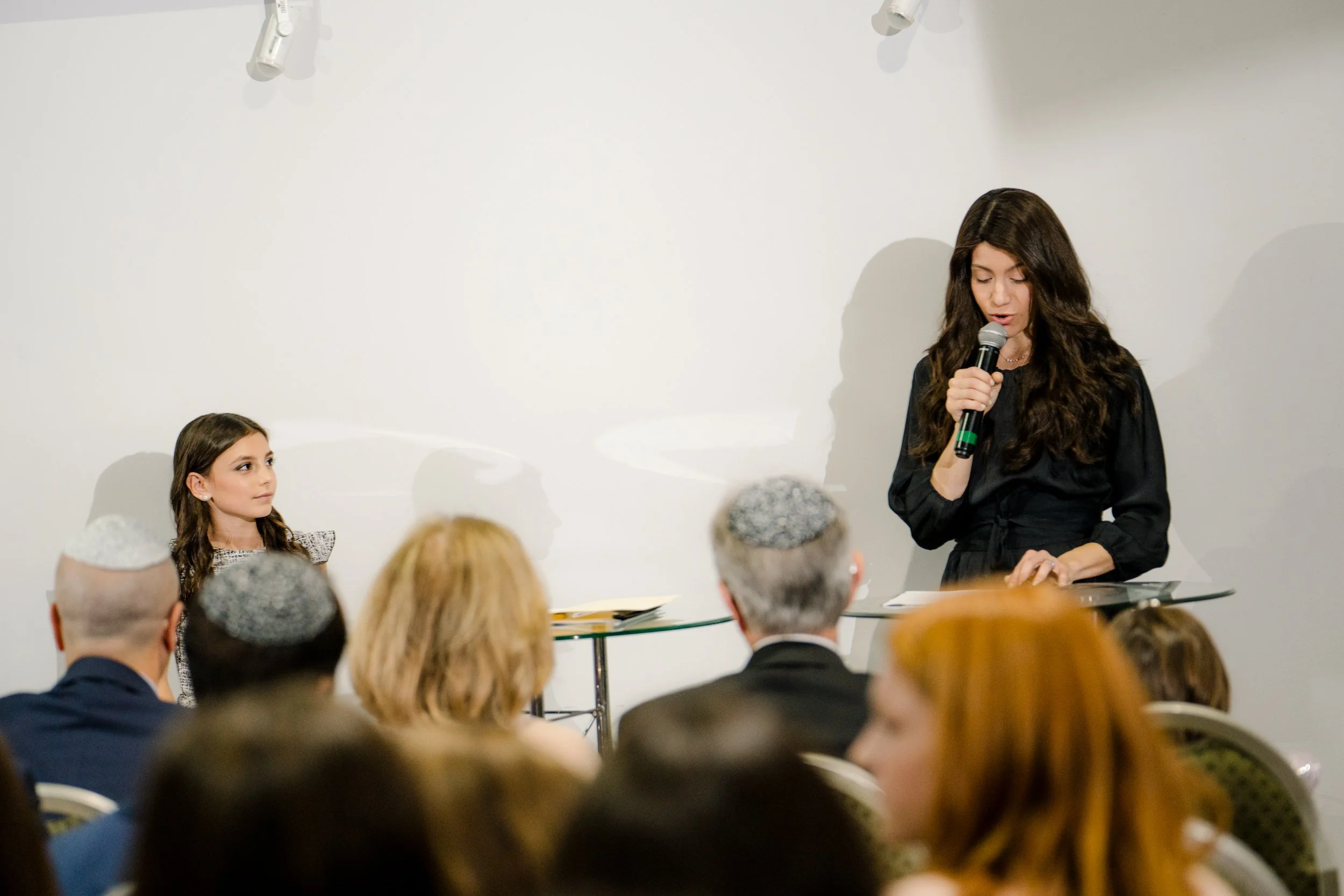 A woman holding a microphone speaks in front of a small audience. A young girl sits at a table on the left. People in the audience are wearing kippahs.