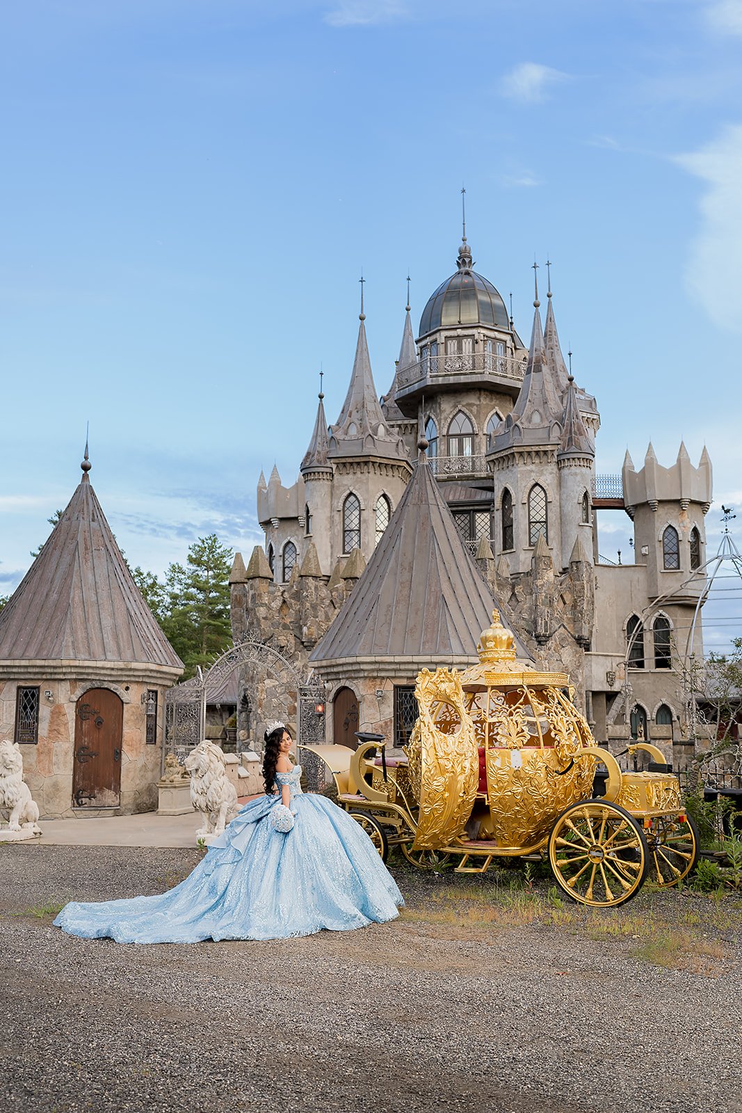 A quinceañera in a blue ball gown standing next to a gold carriage in front of Christ Mark Castle in Woodstock Conneticut.