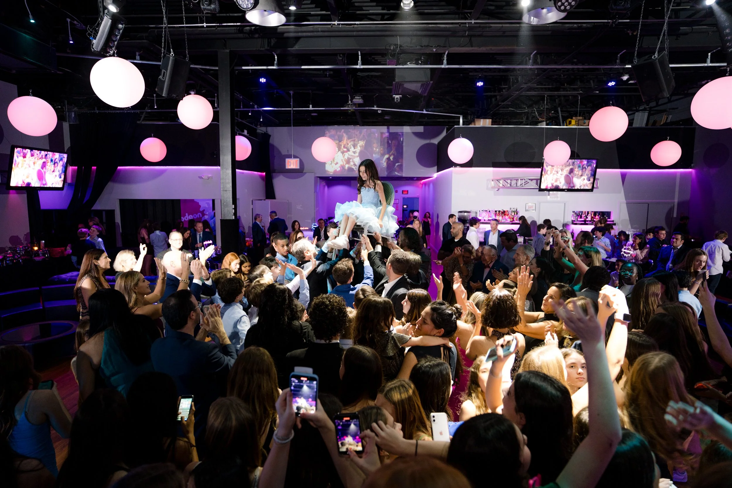 A lively party scene with a group of people celebrating indoors, focusing on a girl in a blue dress being lifted on a chair as part of a traditional dance. The room is decorated with round, pink lights and has a modern design. The crowd around is che