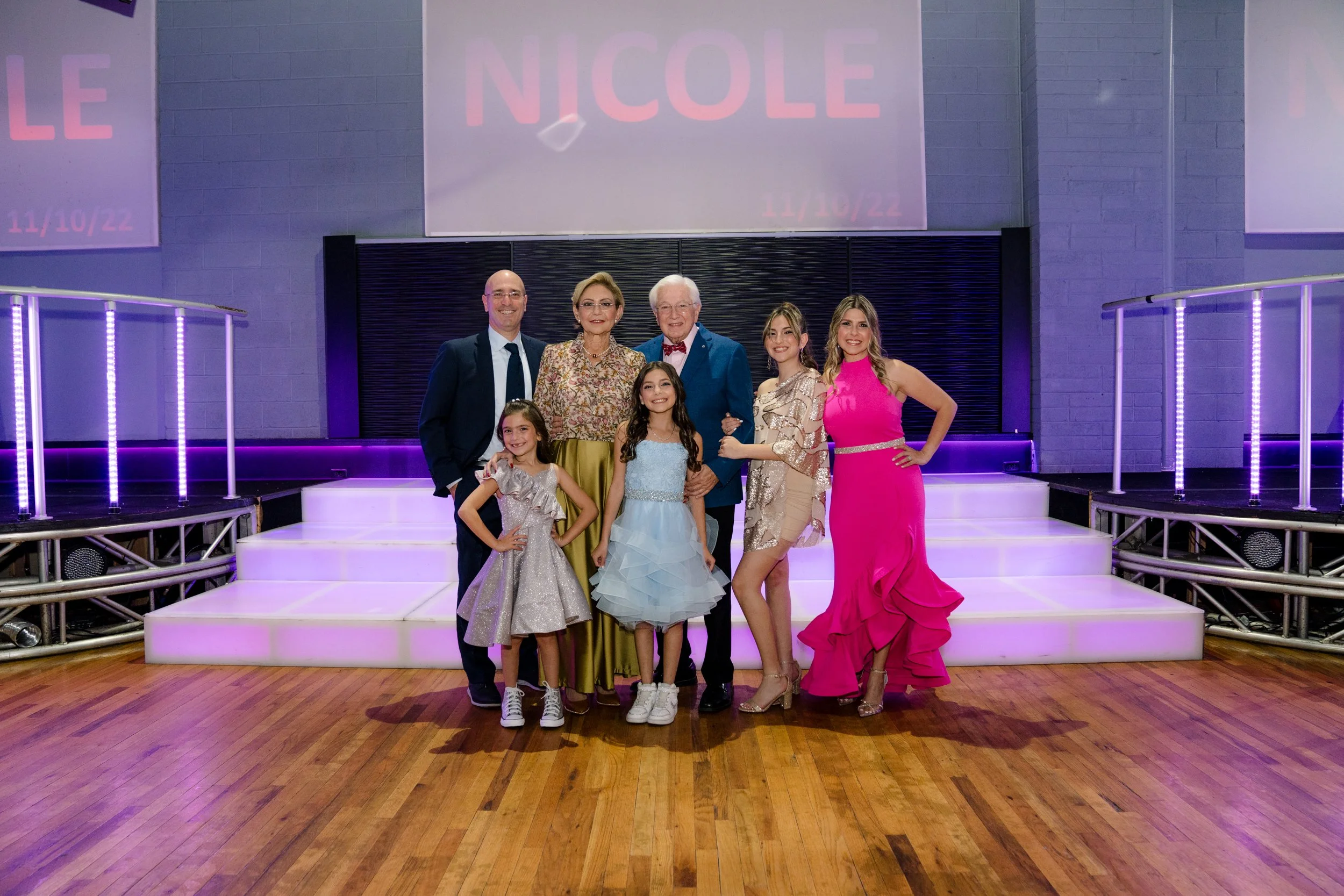 A family poses for a formal photo in a venue with a lit stage and decorative lighting. They are dressed in formal attire, including dresses and suits. The background features a sign with the name "Nicole" and a date "11/10/22.” The setting suggests a