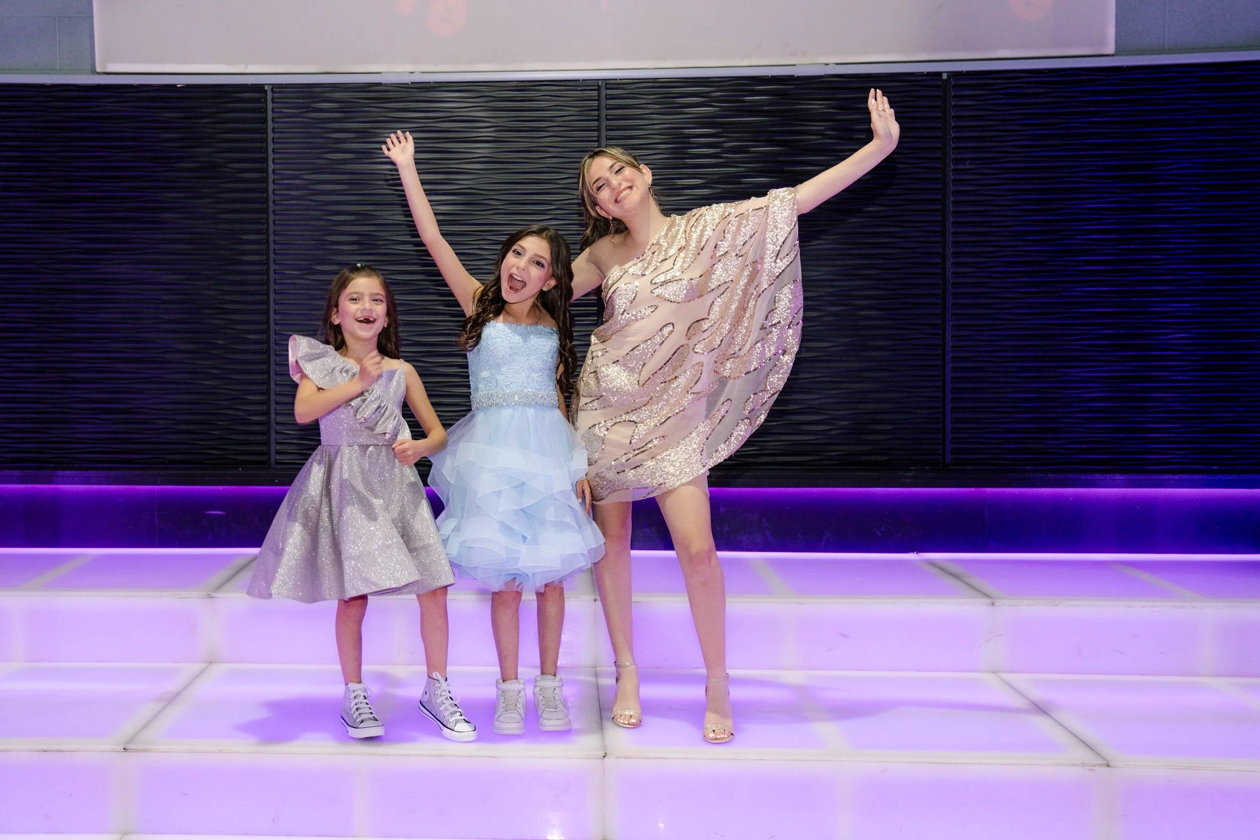 Three girls in stylish dresses posing on a purple-lit floor, with one girl in silver sneakers, another in pastel blue dress, and the tallest in a sequined one-shoulder dress.