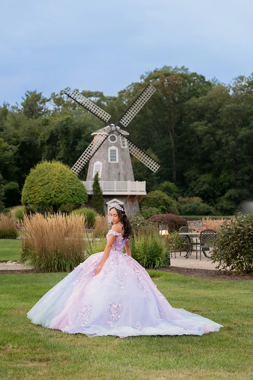 Sweet sixteen girl in a lavender ball gown standing outdoors near a traditional windmill with lush green trees in the background.
