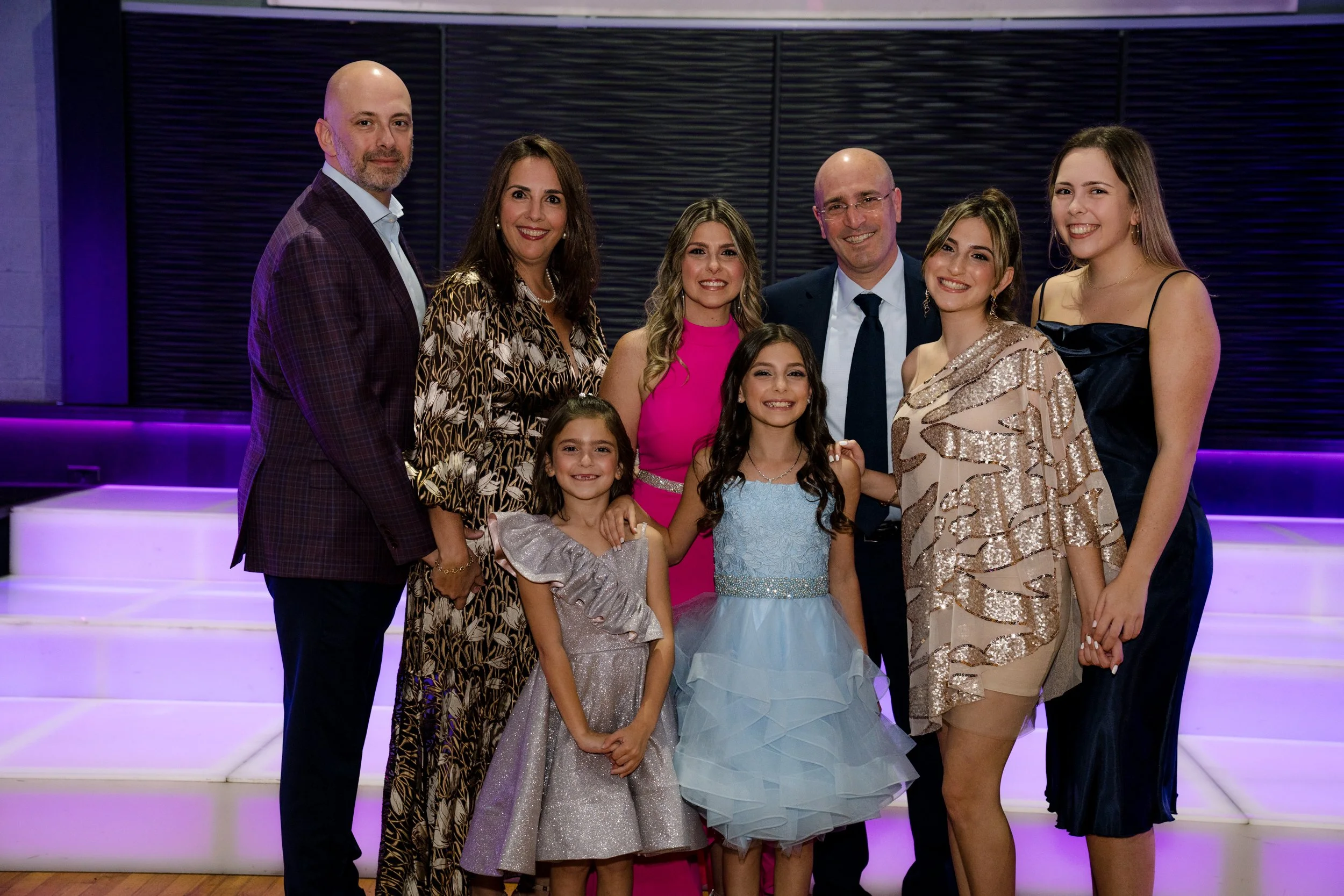 A group of smiling people posing together in formal attire at a party or event. The background has illuminated stairs with a purple hue.