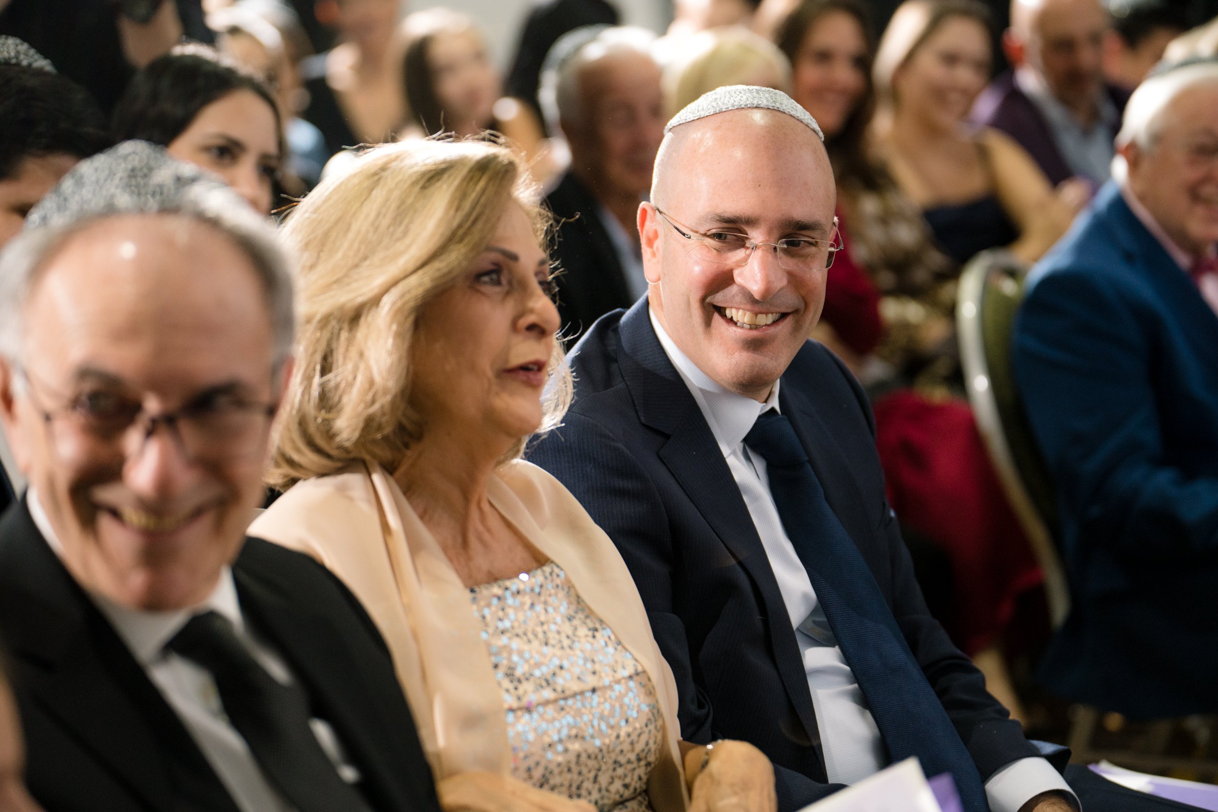 A group of people sitting at an event, including a man in a suit with glasses and a kippah, smiling at a woman with shoulder-length blonde hair wearing a sparkling dress, among others in the background.