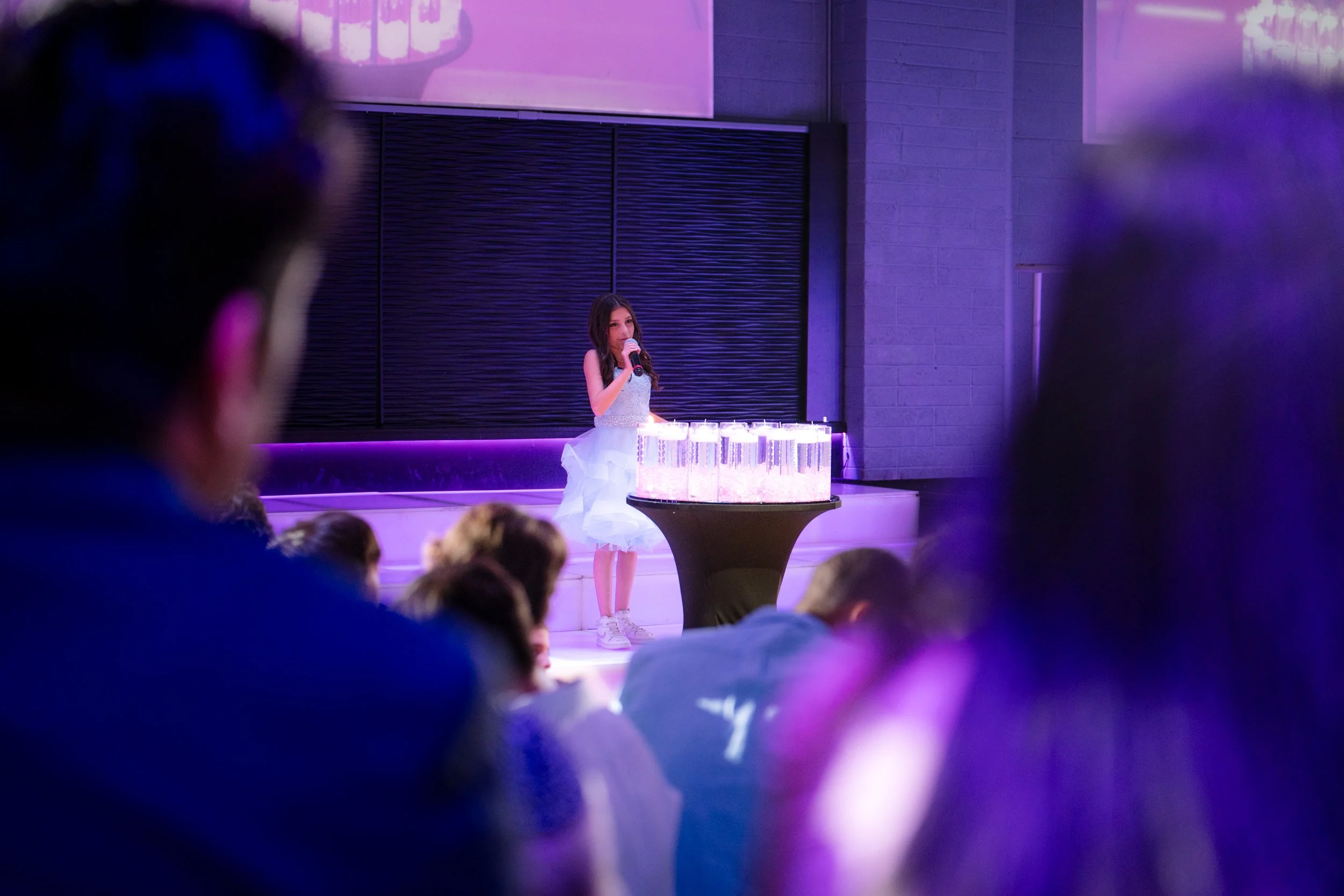 A young girl in a light blue dress speaking into a microphone on stage with a table of clear cylindrical decorations, surrounded by an audience in a dimly lit room with purple lighting.