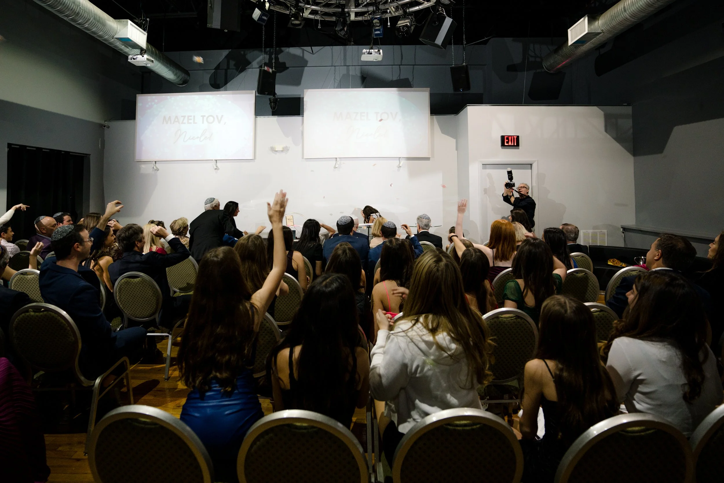 Group of people in a room attending a celebration, with a screen displaying 'Mazel Tov' and a photographer capturing the event.