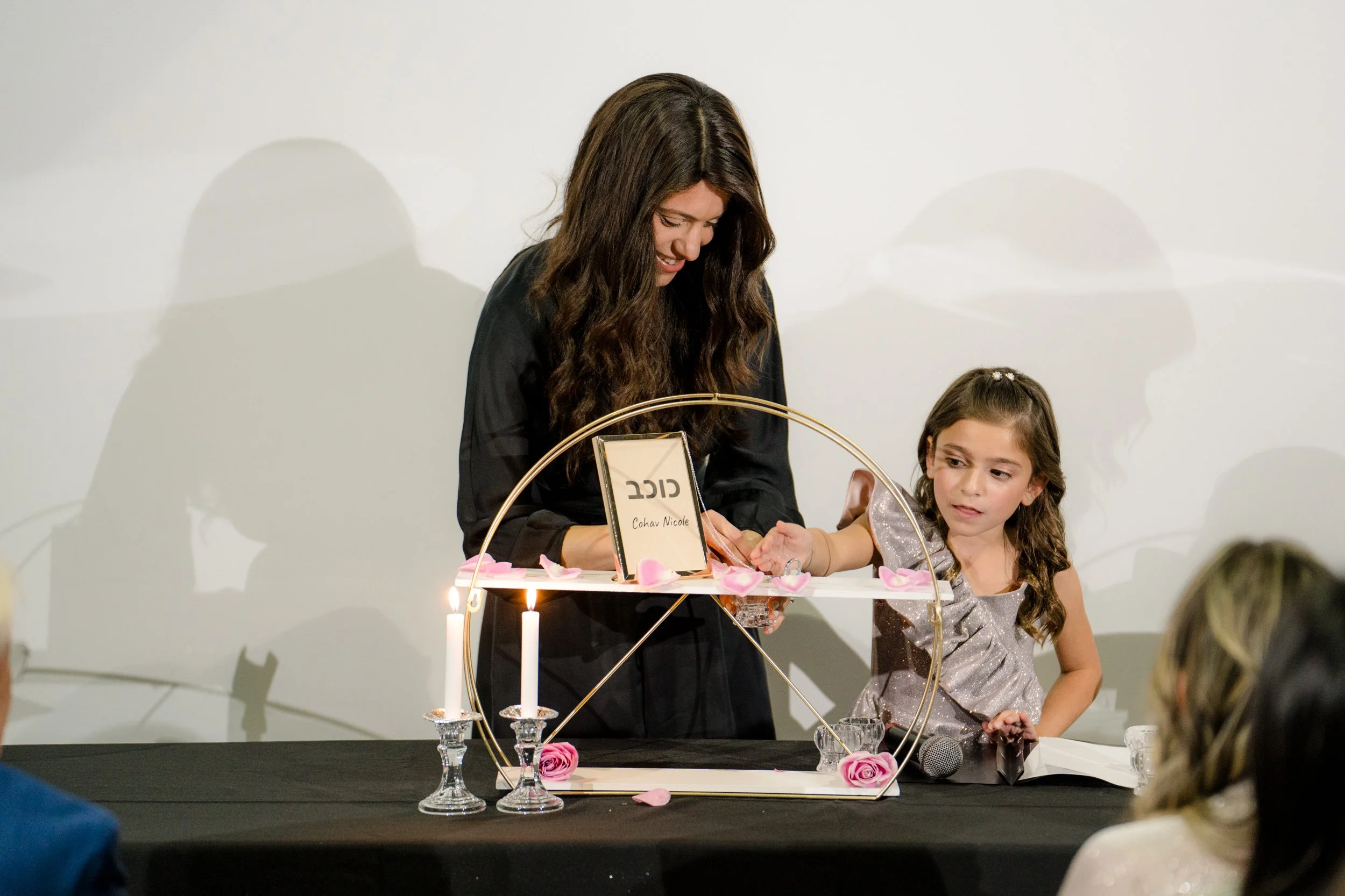 Woman and girl lighting candles on a table adorned with pink roses and petals, featuring a framed nameplate with Hebrew text, against a neutral background.