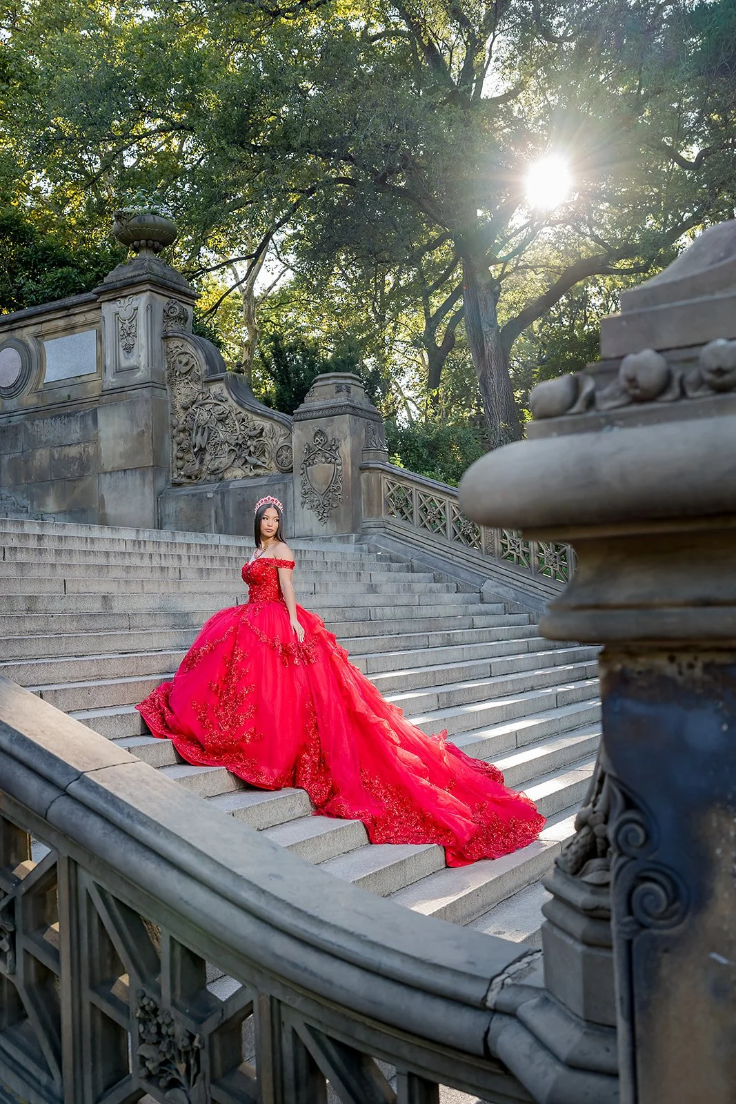 A Sweet sixteen girl in a bright red, elaborate gown with floral details, standing on the stone stairs of Bethesda Terrace at Central Park, with ornate stone railings and trees in the background, sunlight shining through the branches.