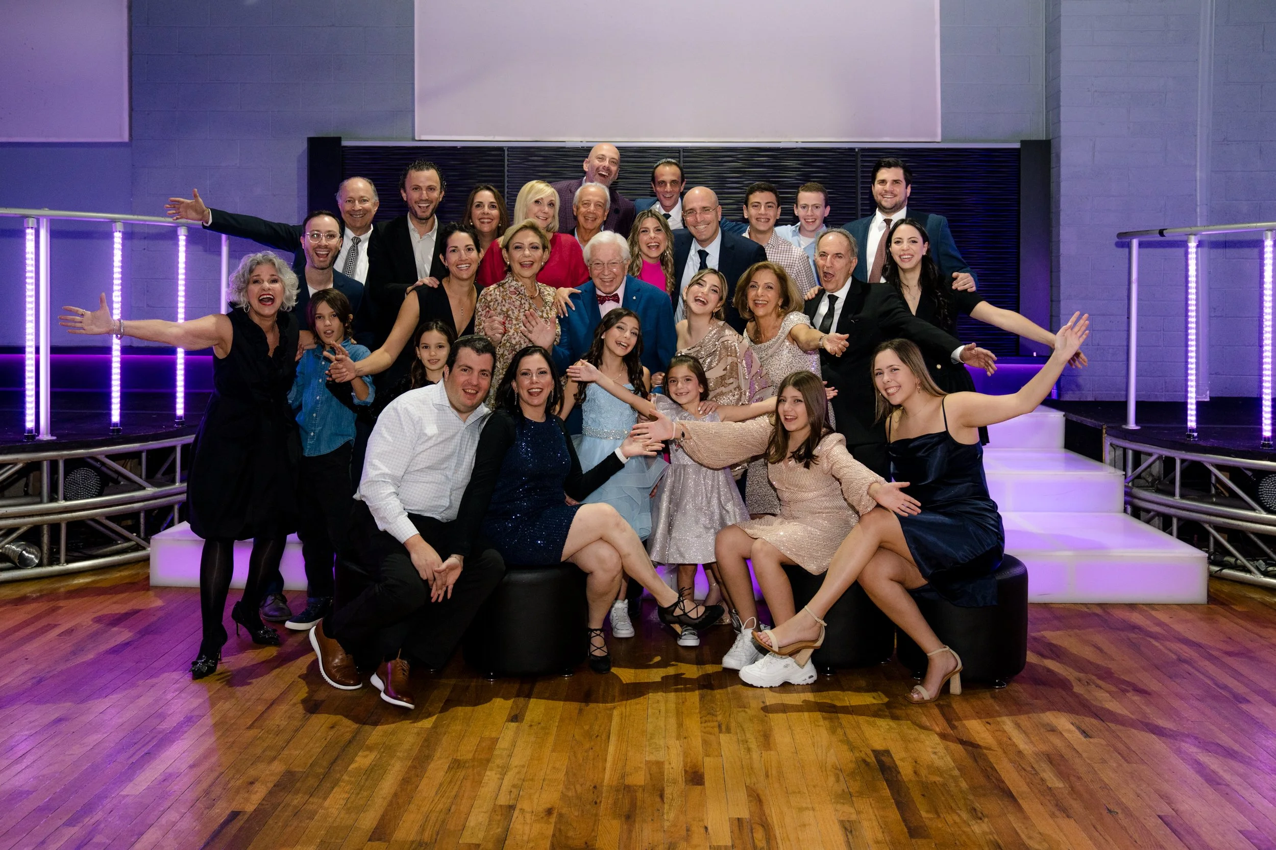 A large group of people posing together at an indoor event with a party atmosphere, featuring purple lighting and a wooden floor. They are smiling and appear to be in a celebratory mood.