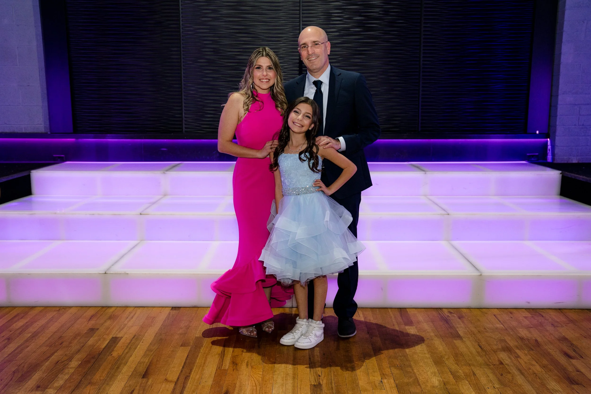 A family of three posing on a wooden floor in front of glowing purple steps. The woman is wearing a bright pink dress, the man is in a dark suit, and the young girl is in a light blue dress with white sneakers.