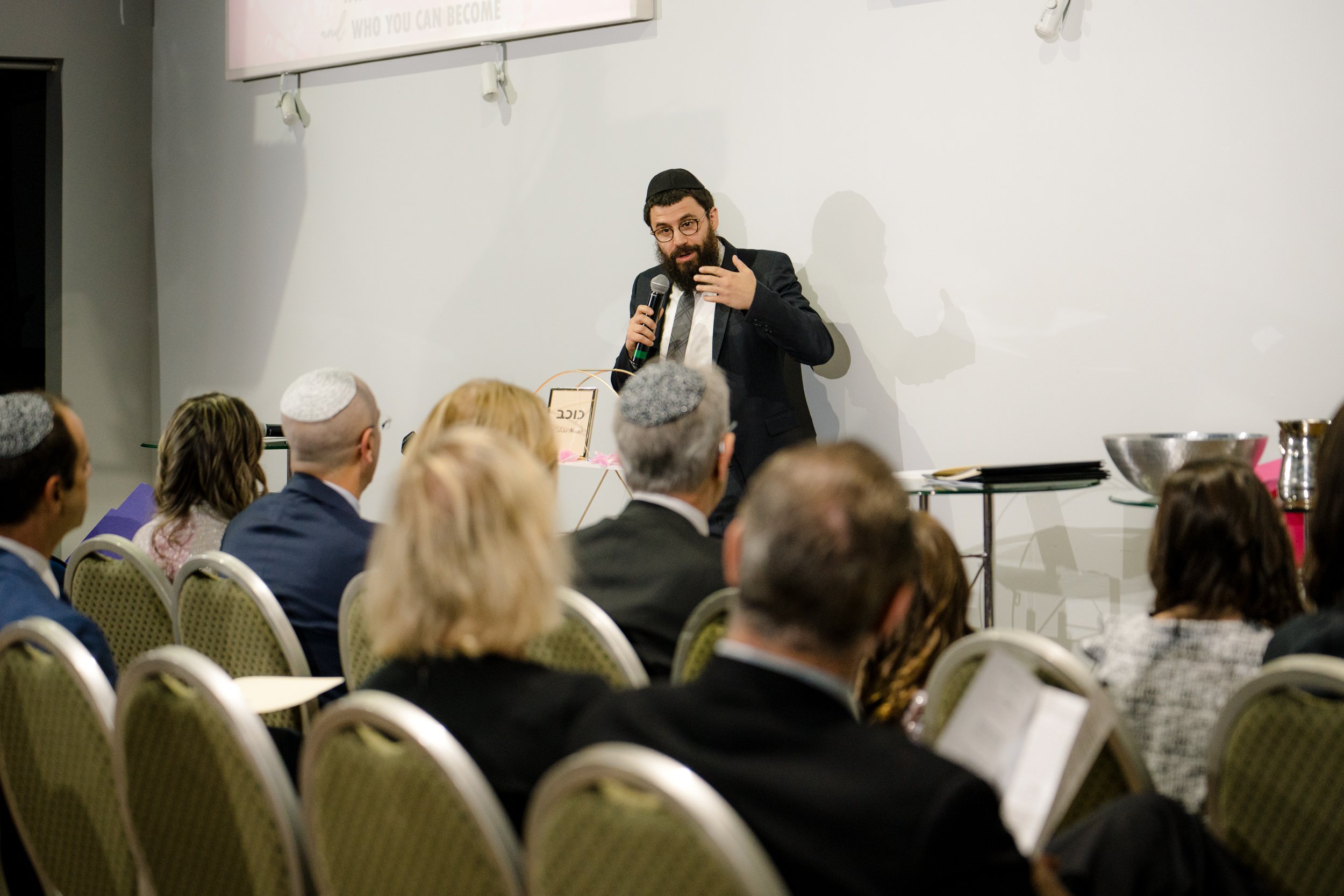 A man speaking to an audience, holding a microphone, wearing a suit and a kippah, standing in front of a white wall. The audience is seated in rows, some wearing kippahs.
