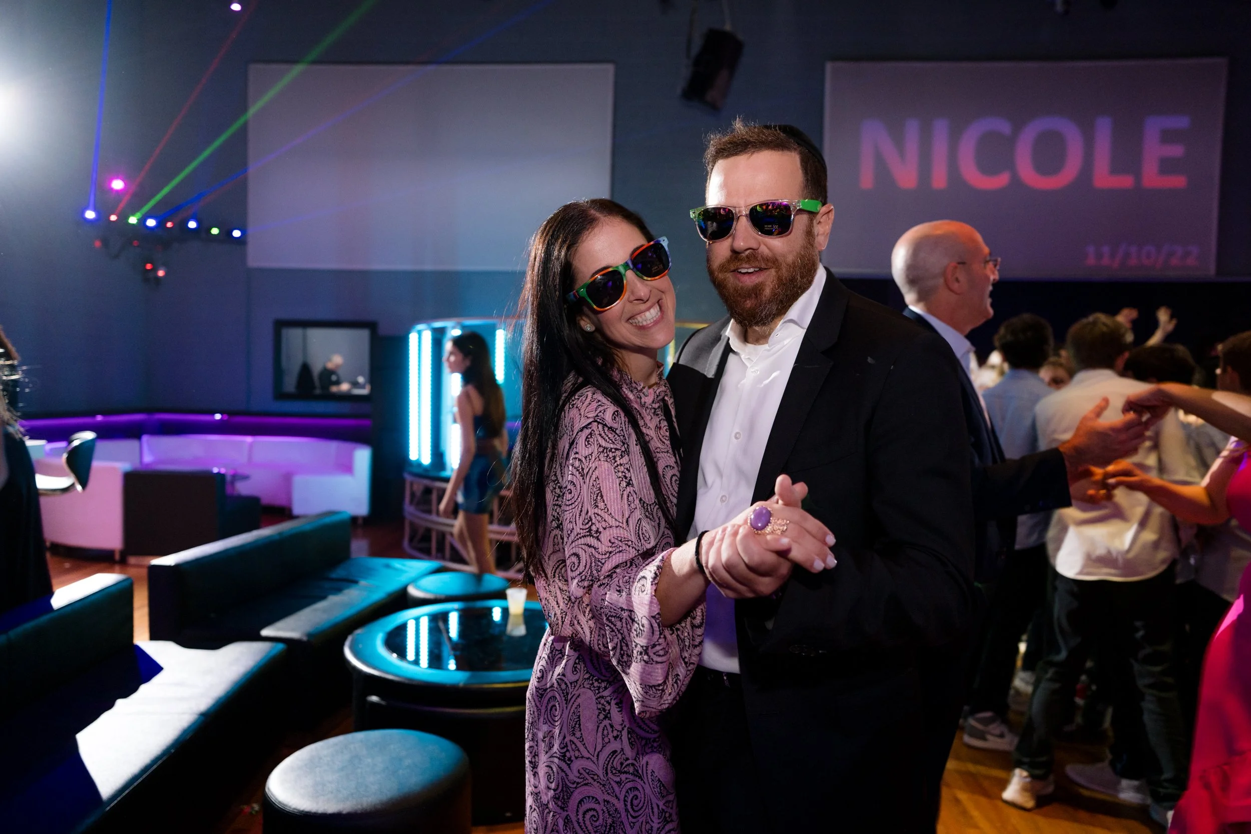 Smiling couple wearing sunglasses dancing at a party with colorful lights and people in the background.
