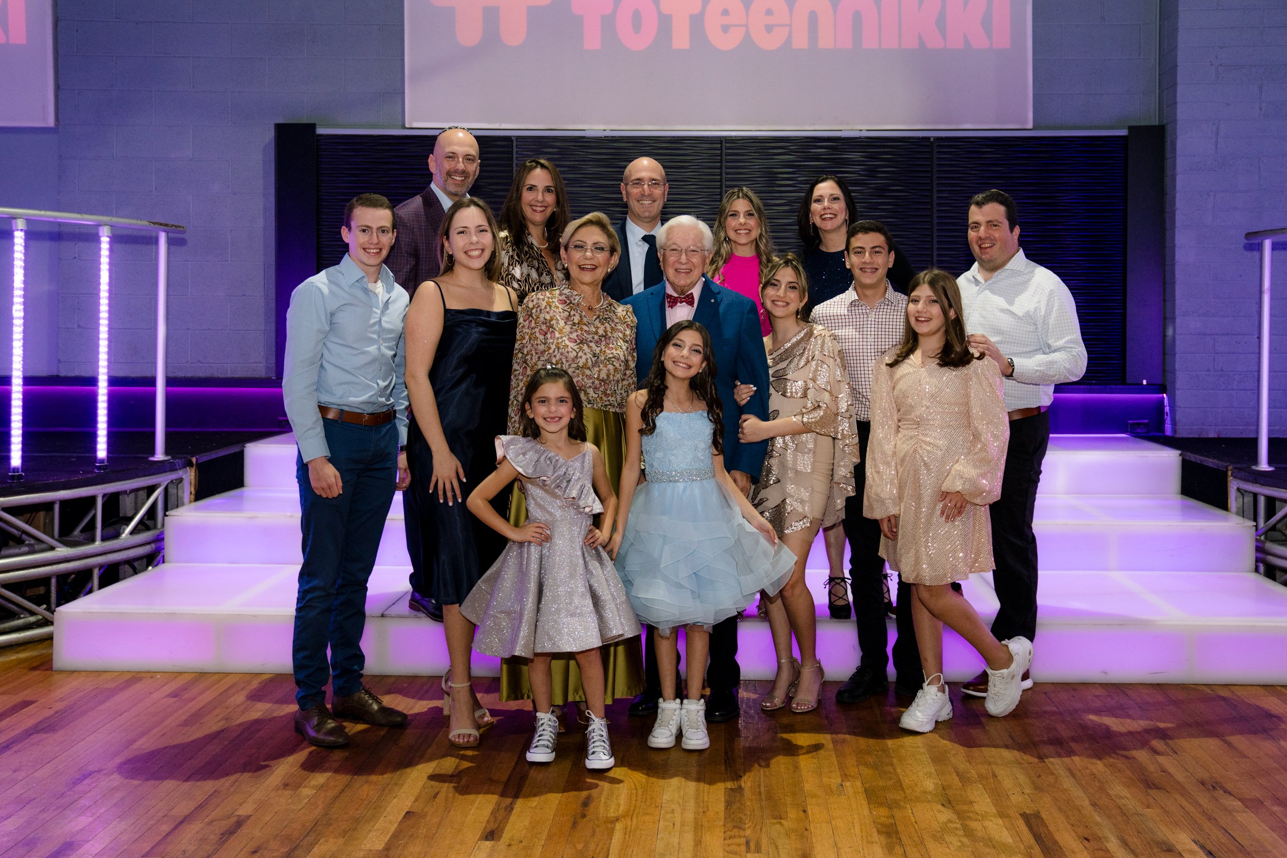 Group of people posing on a dance floor with purple lights, featuring men, women, and children dressed in formal attire, against a backdrop with 'Toteennikki' sign.