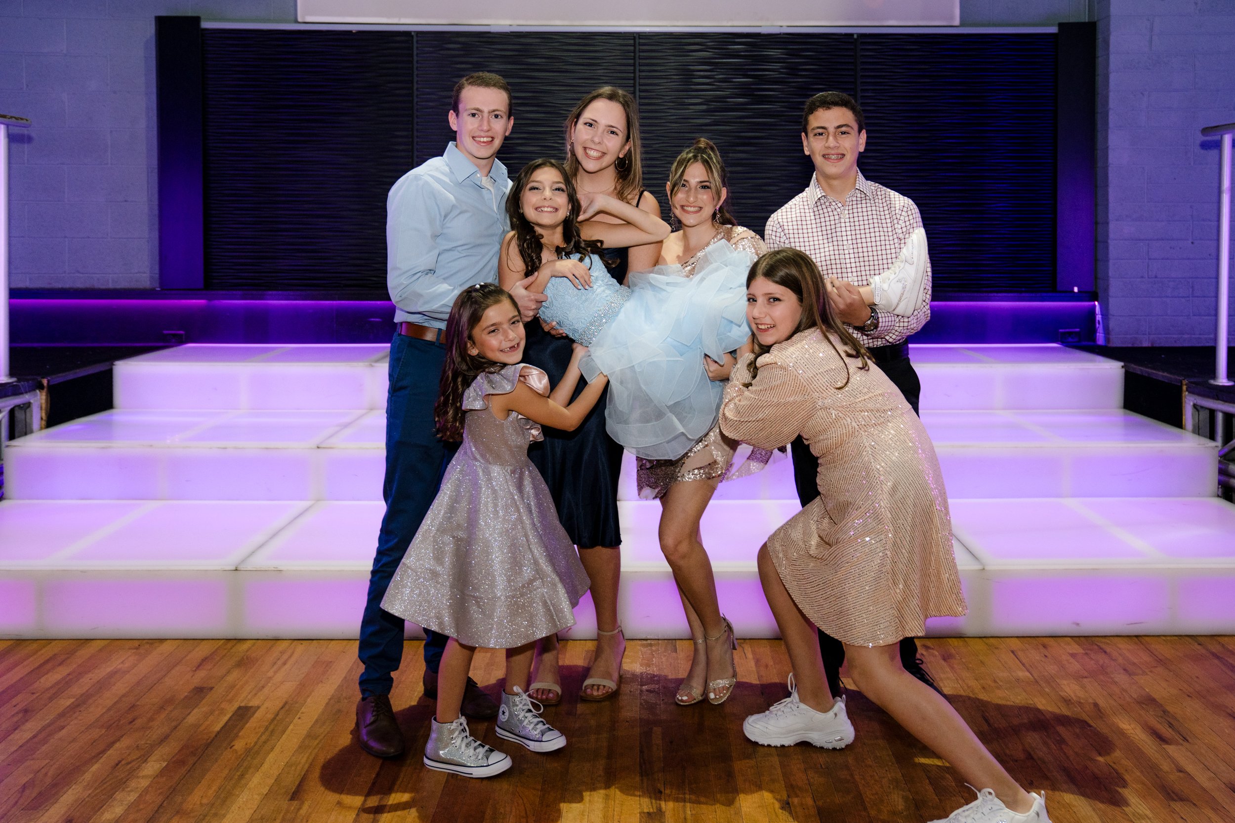 Group of seven people posing indoors, including children and teenagers, with one person being held up by the others. They're standing on a wooden floor in front of a raised platform with purple lighting. Everyone is dressed in formal or festive attir