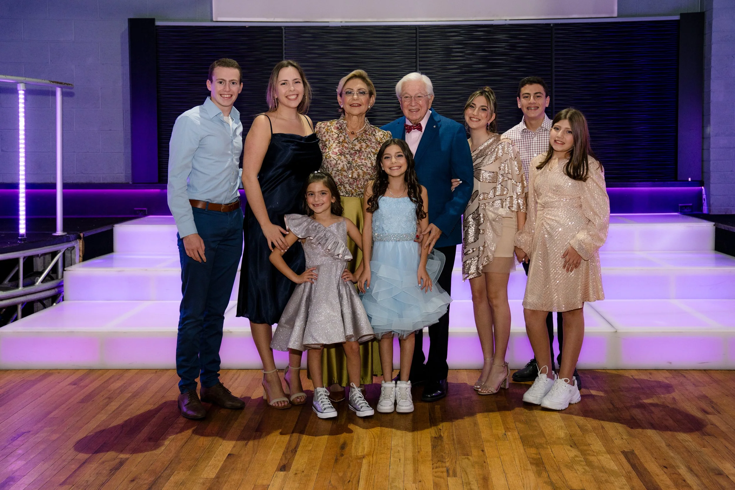 Group of people dressed formally posing on a wooden floor with lit steps and a dark backdrop.
