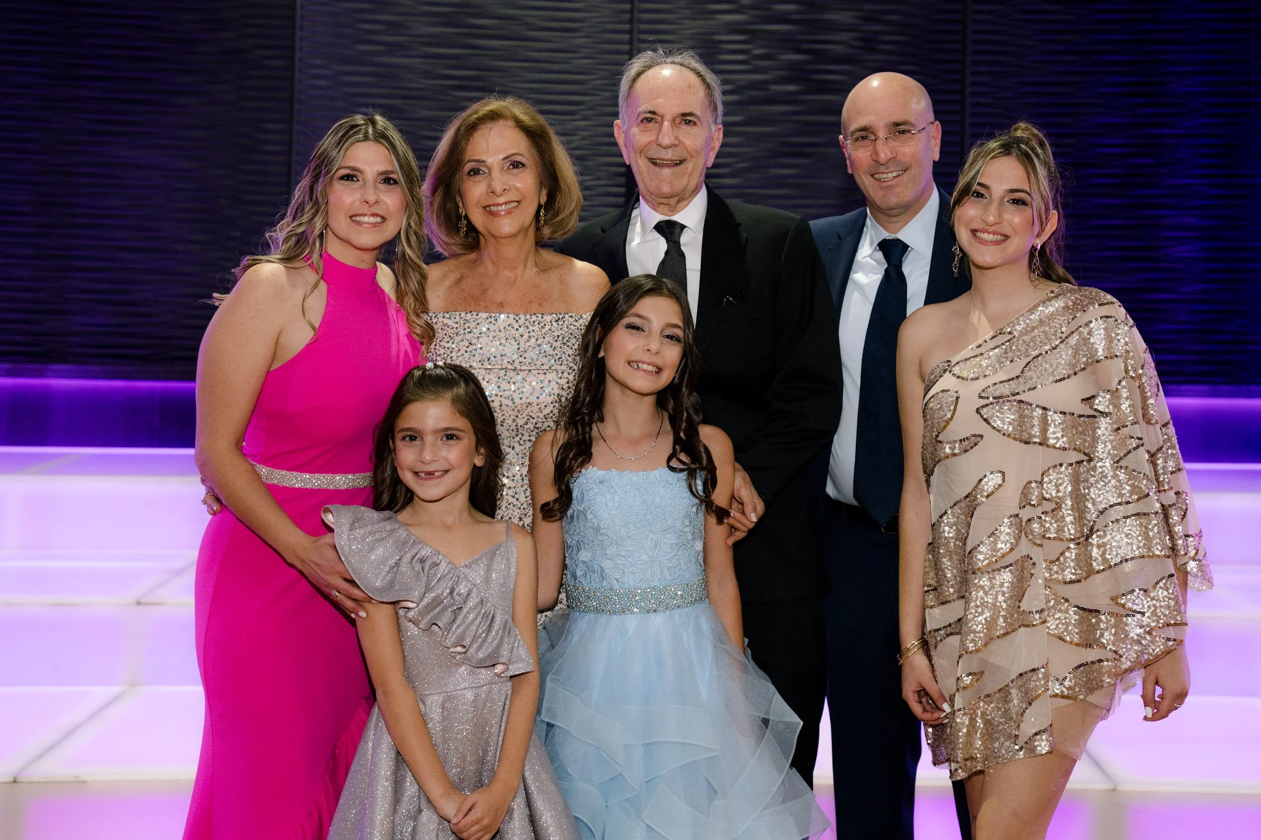 A group of seven people dressed formally, posing together in front of a purple-lit background. There are four adults and three children, all smiling.