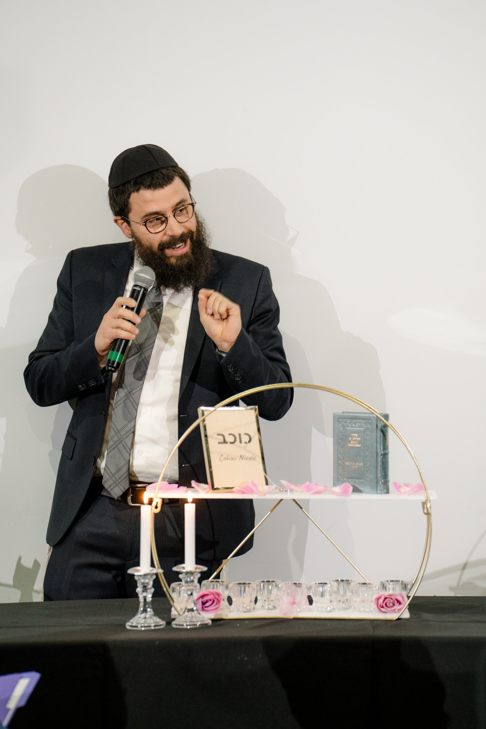 Man in a suit and kippah speaking into a microphone beside a decorated table with candles and religious items.