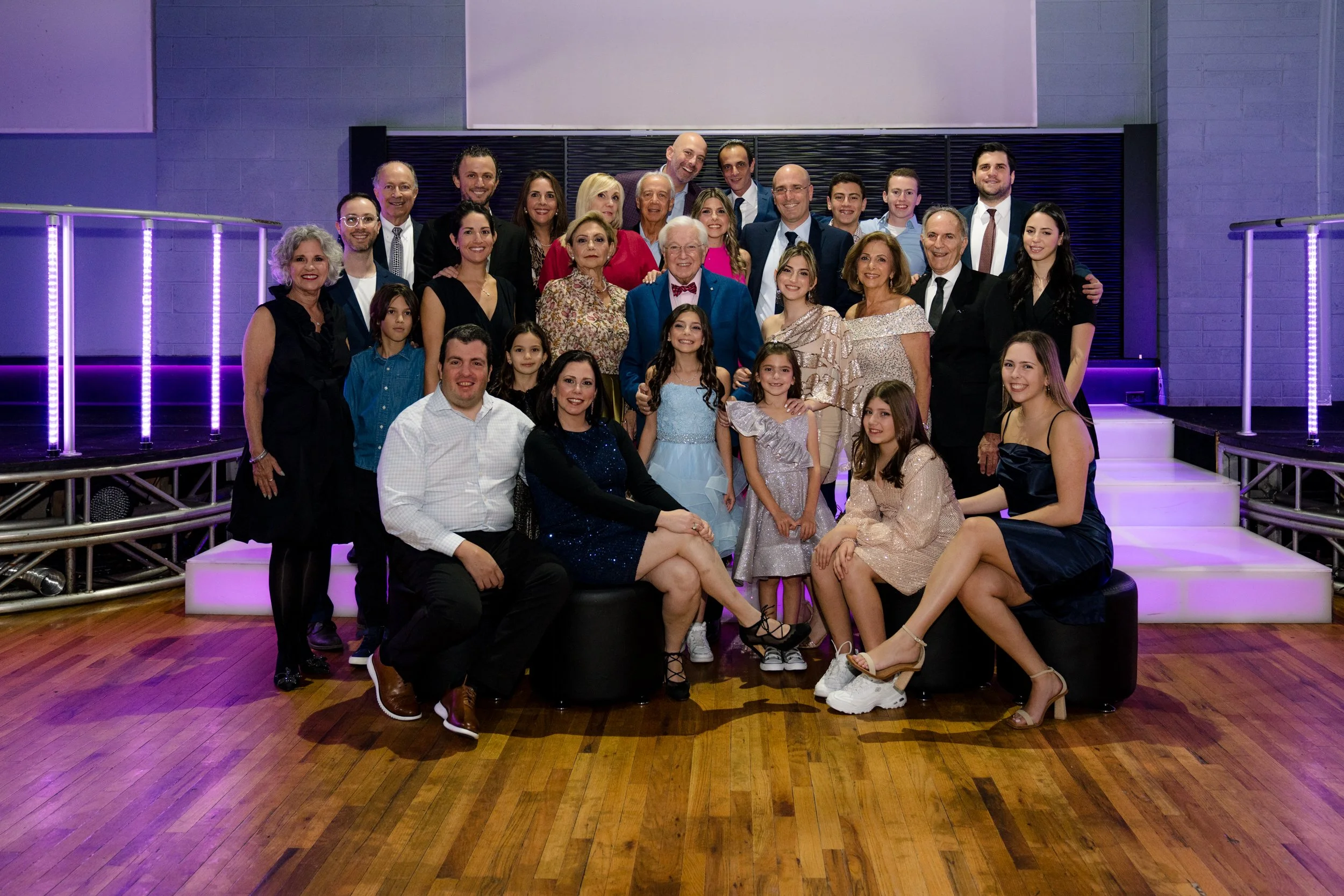 A large group of people posing together at an indoor event, with women, men, and children dressed in formal attire, smiling, standing and seated on a wooden floor with purple lighting in the background.