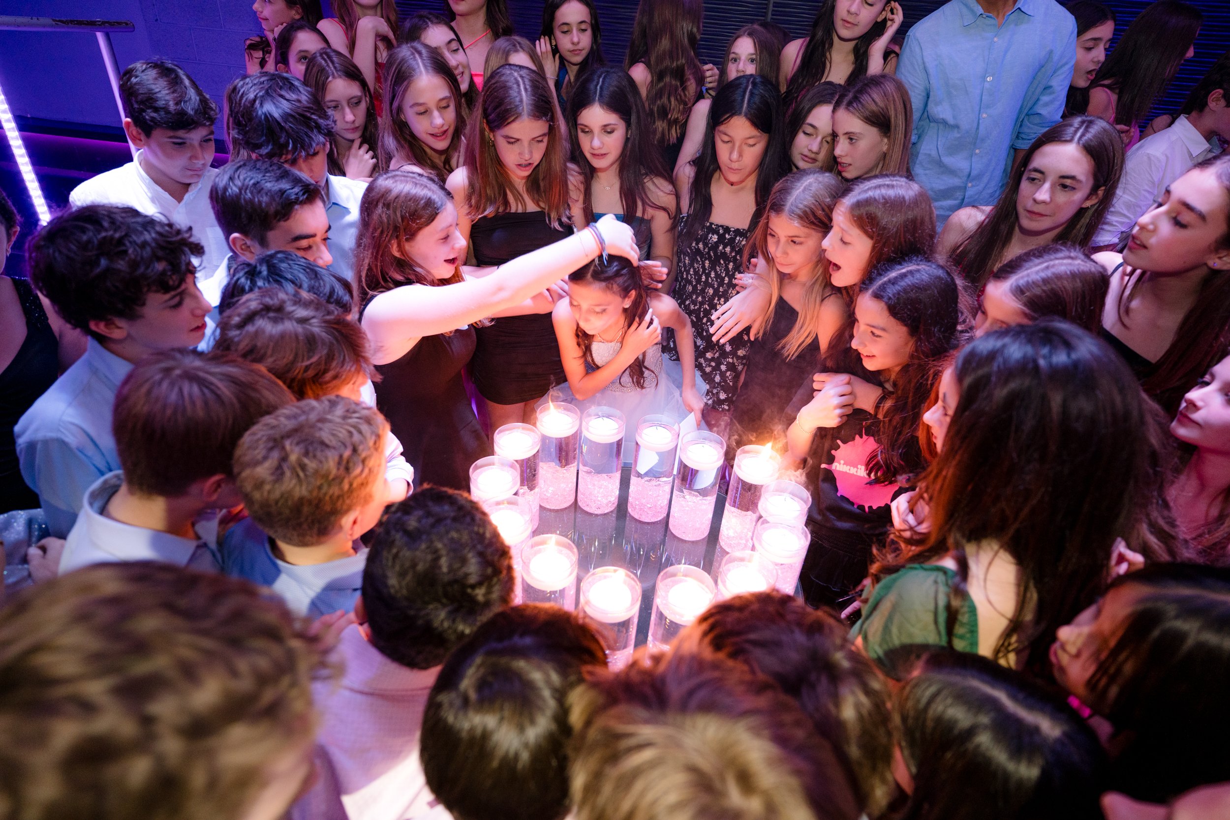 Group of people standing around a table with lit candles, celebrating an event in a dimly lit room.