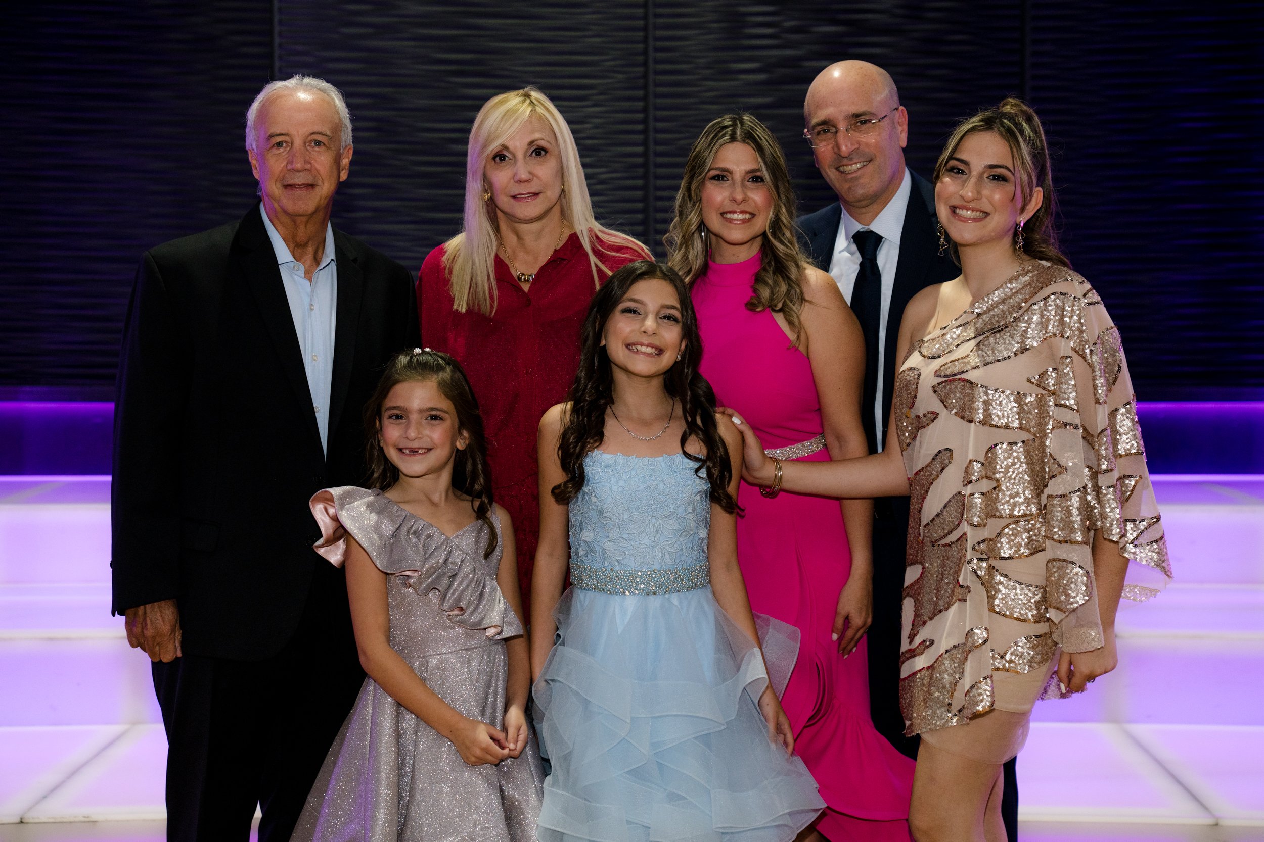 A family group photo with three adults and four children, all well-dressed in formal attire, standing on a lit staircase with a dark backdrop and purple lighting.