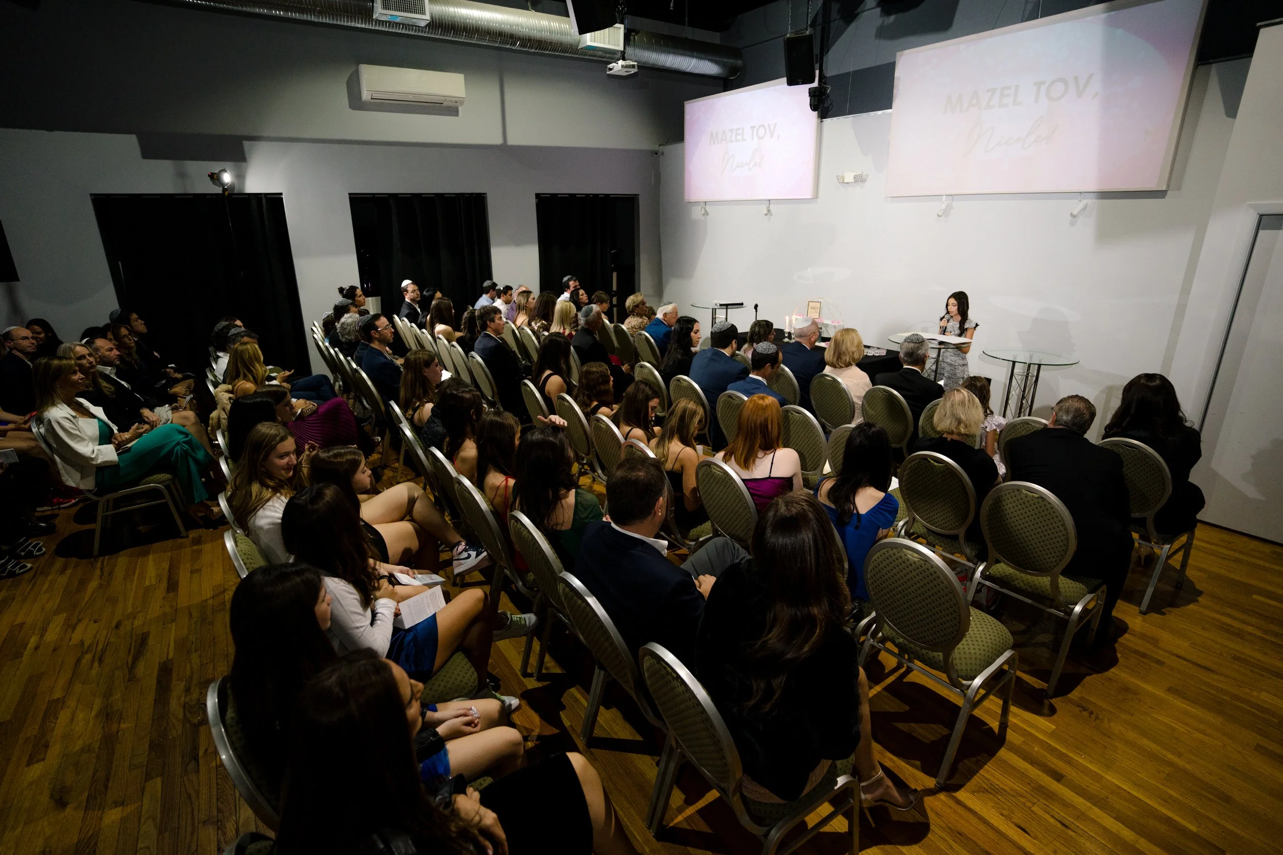 Audience attending an indoor event with a speaker at the front and screens displaying 'Mazel Tov'