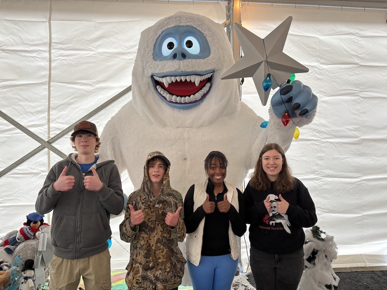 Four people posing in front of a large Yeti figure holding a star and string lights. They are inside a tent with winter decorations, including snowmen figures.