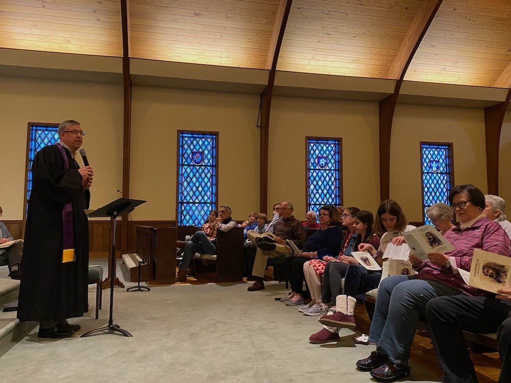 A minister speaking at a church service with a microphone, standing beside a podium. Congregation seated in pews with stained glass windows in the background.