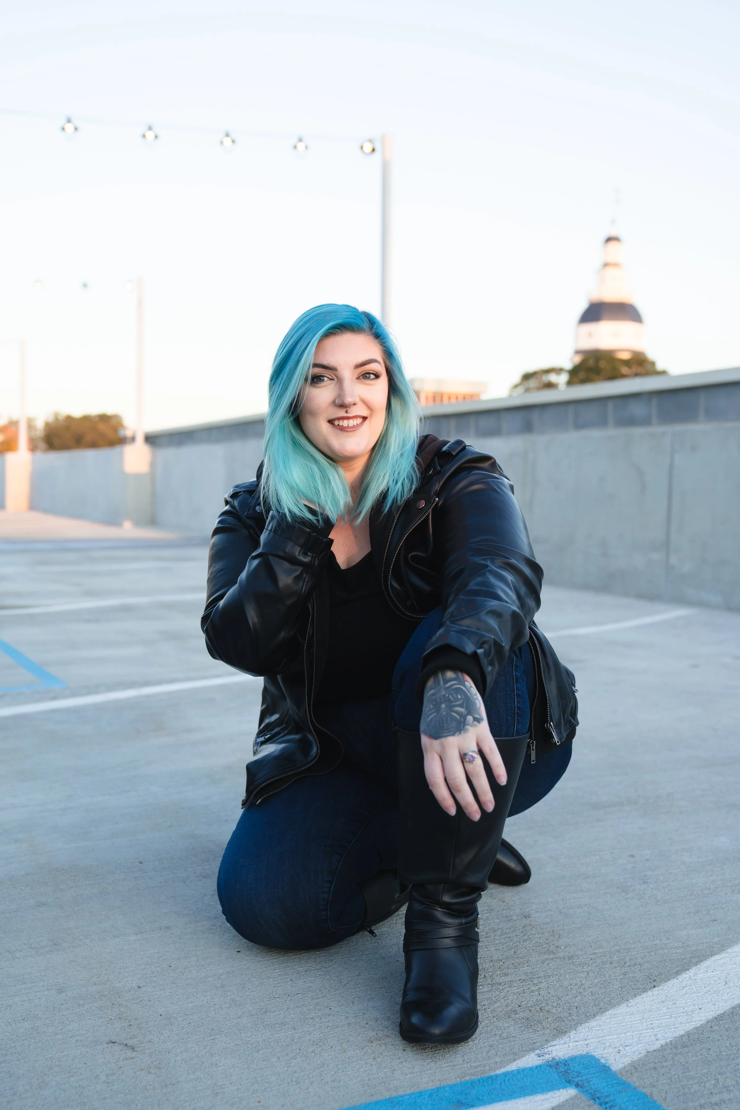 A woman with turquoise hair, wearing a black leather jacket and black boots, is crouching on a rooftop parking lot during sunset, smiling at the camera.