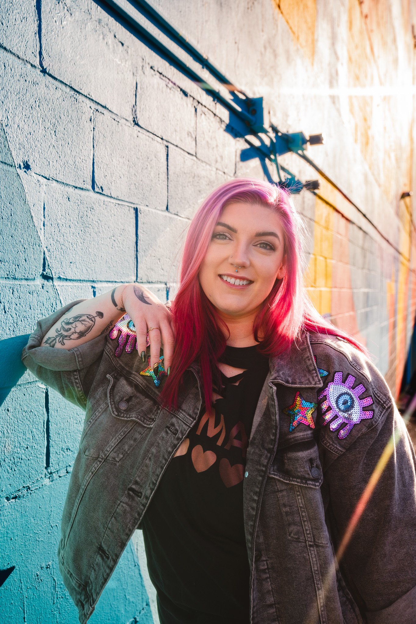 A young woman with pink hair leaning against a colorful painted brick wall, smiling at the camera, wearing a denim jacket with patches, a black t-shirt with hearts, and visible tattoos on her arm.