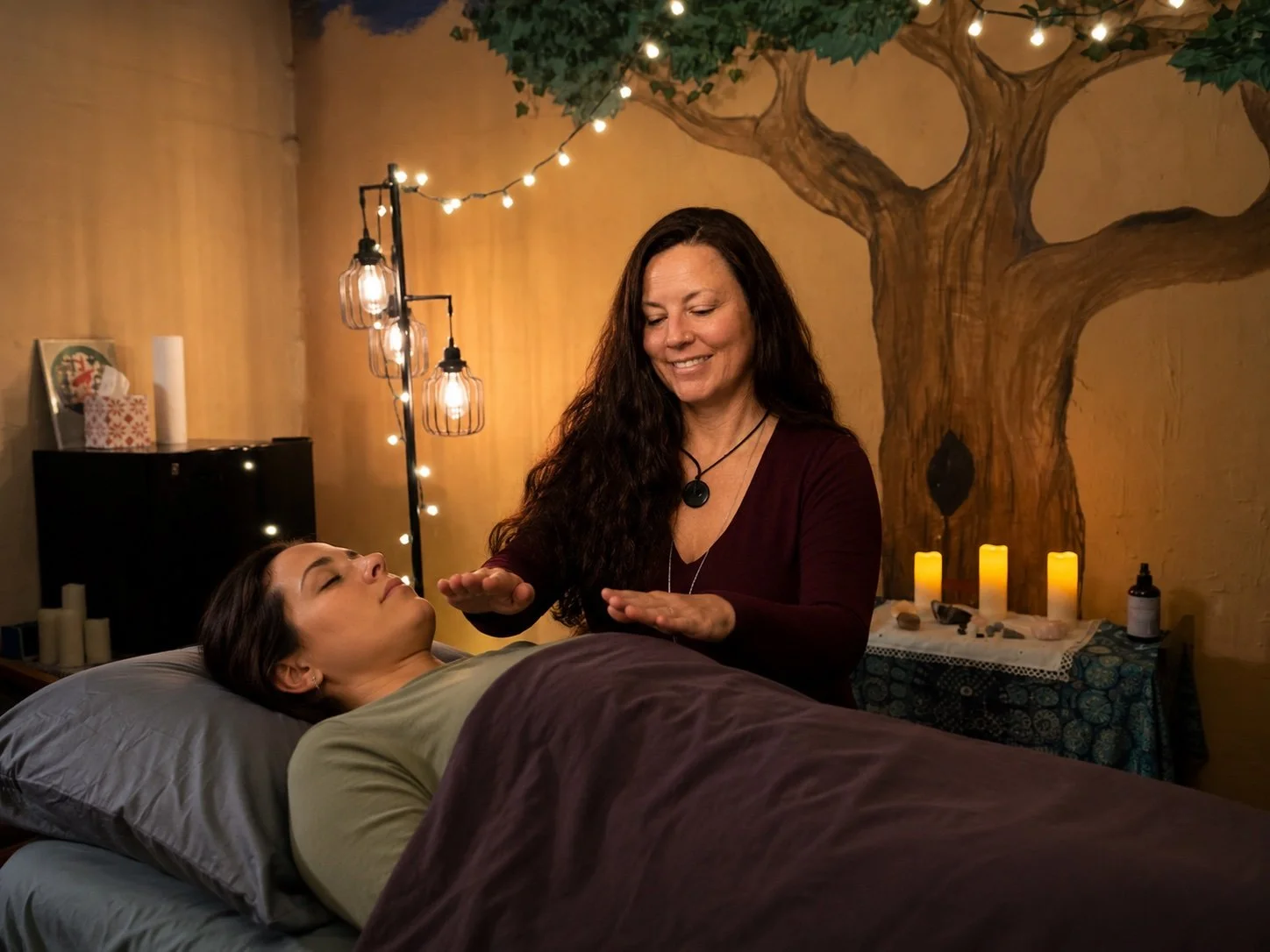 A woman receiving a Reiki healing session from a practitioner in a cozy room with warm lighting, candles, string lights, and a painted tree on the wall.
