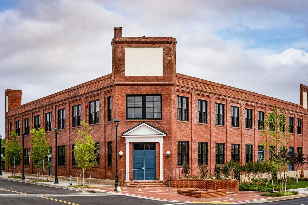 Red brick building with black-framed windows and a blue door, landscaped sidewalk with young trees and street lamps.