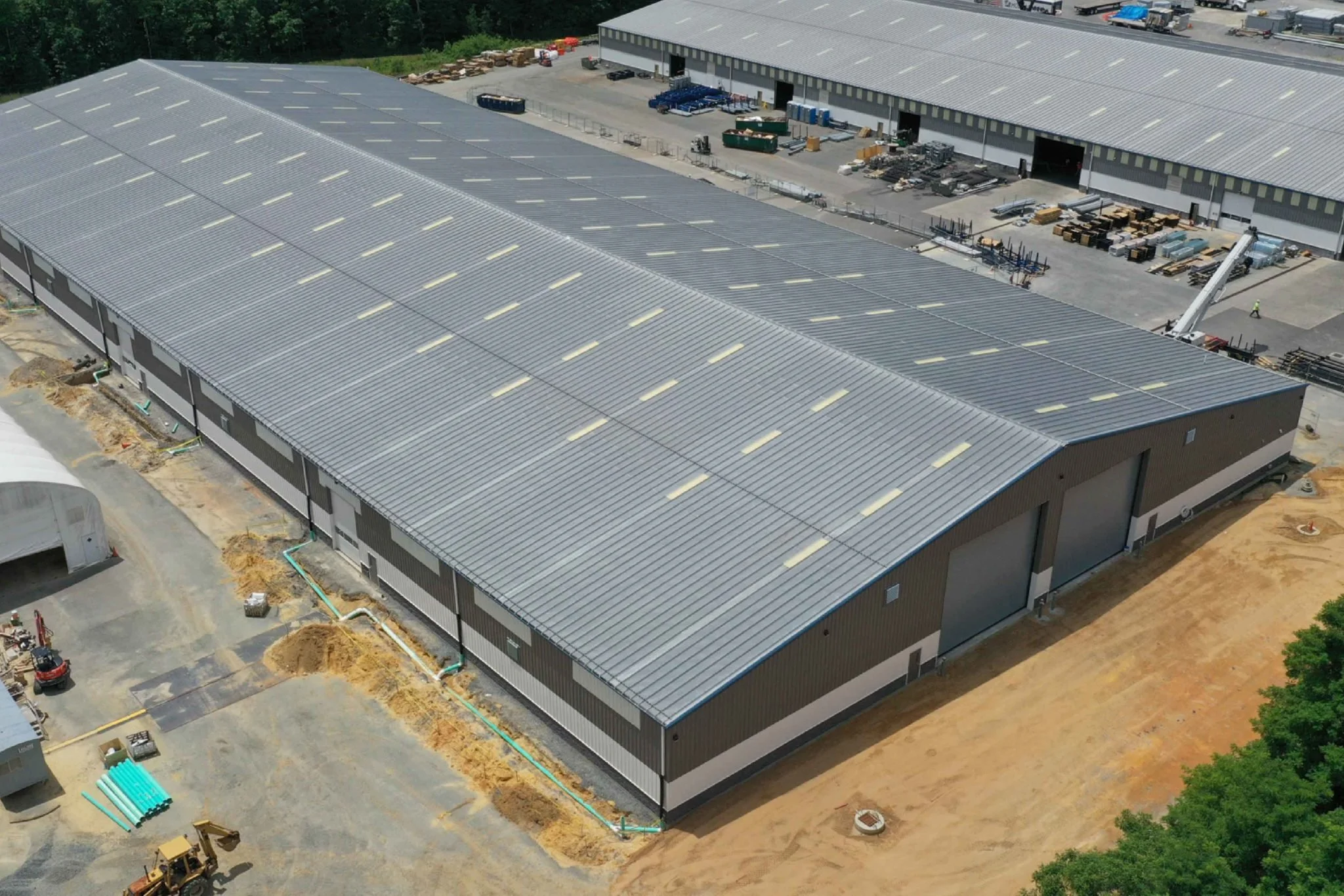 Aerial view of an industrial warehouse under construction with metal roofing and outdoor equipment, surrounded by trees and construction materials.
