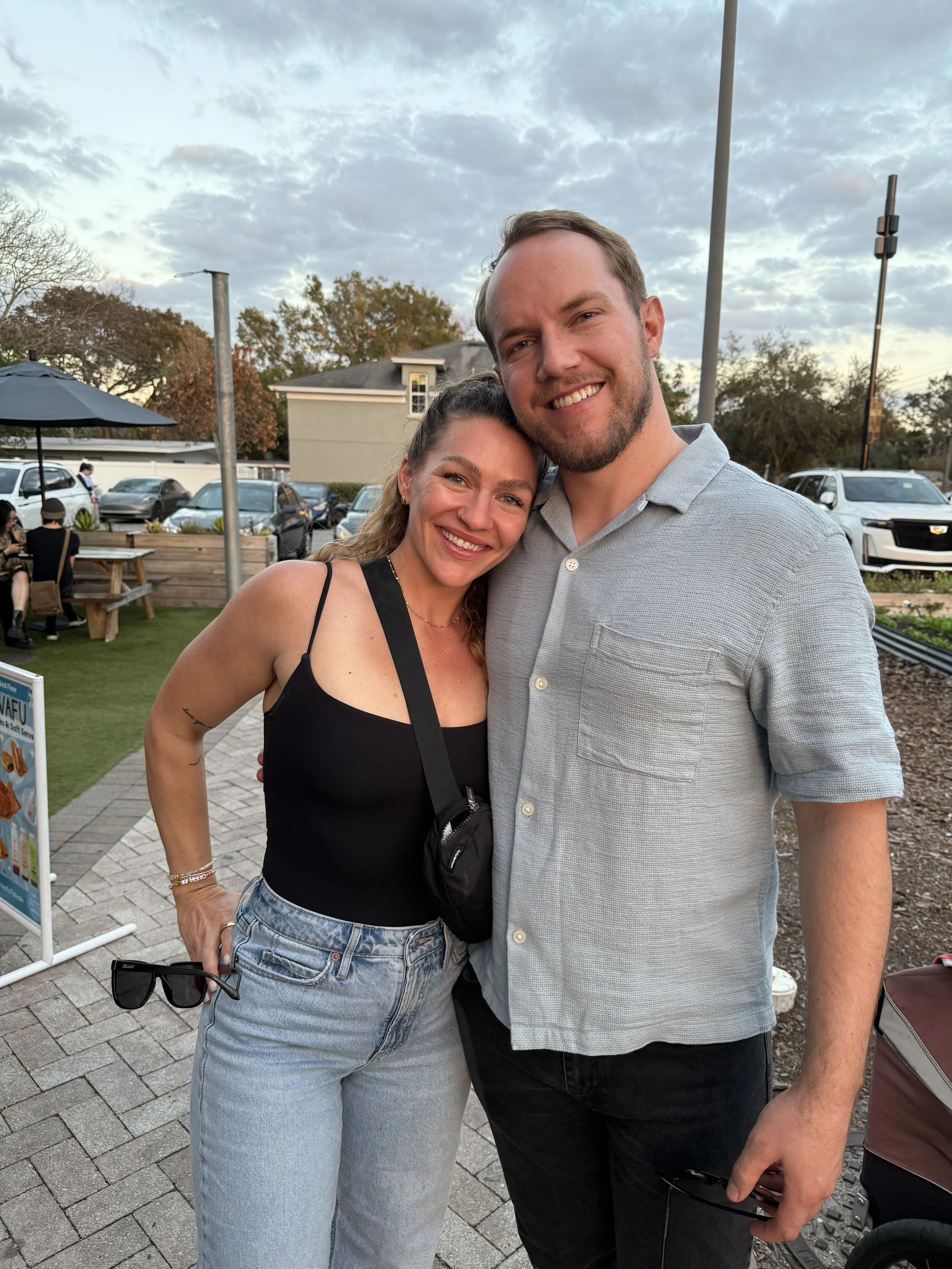 A smiling woman and man pose together outdoors during daytime, with the woman holding sunglasses in her left hand, both dressed casually, standing on a paved walkway surrounded by parked cars, trees, and a small crowd in the background.