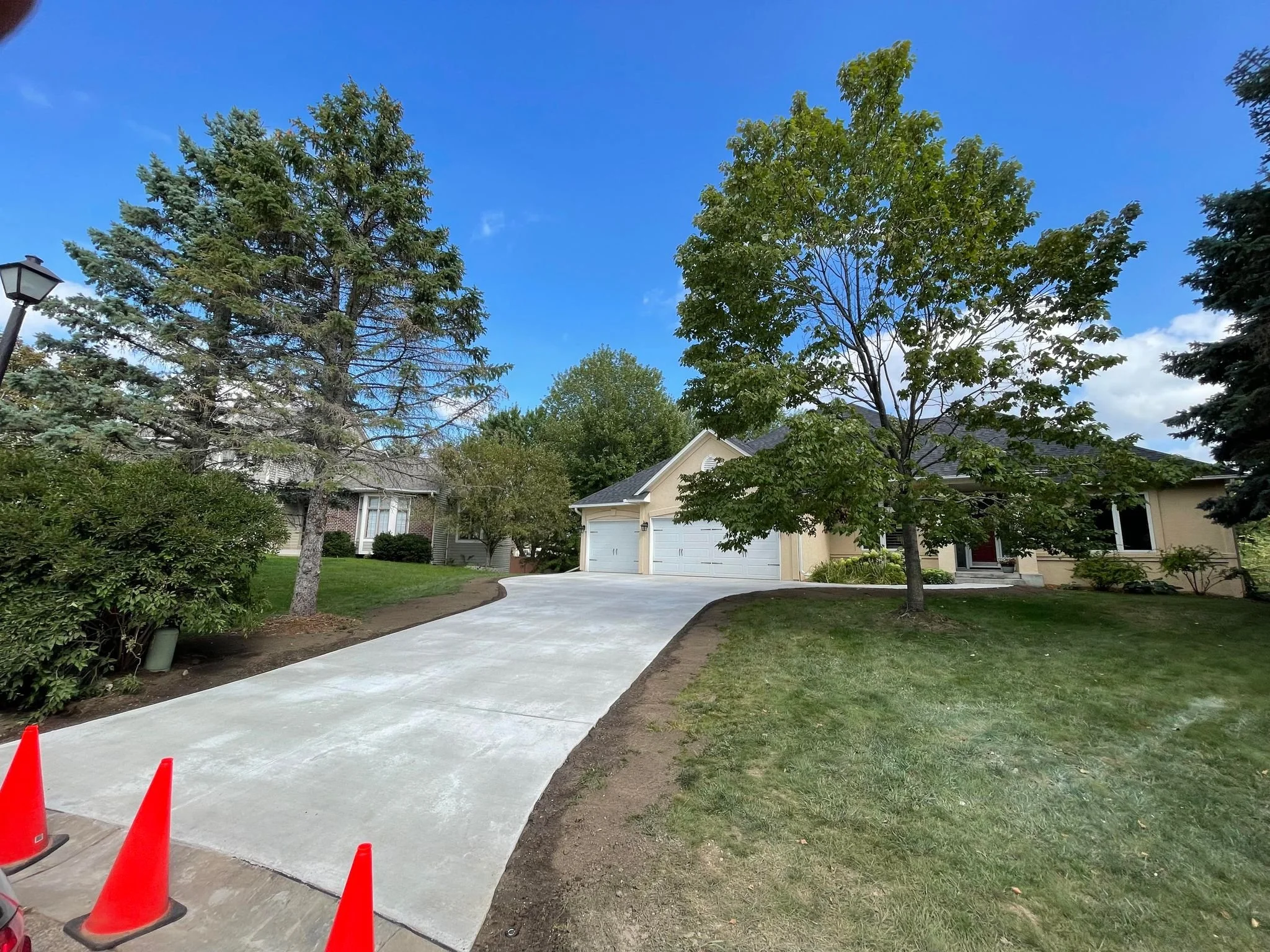 Newly paved concrete driveway in Minnesota leading to a house with a garage. Green grass and trees on either side, a street lamp on the left, and a clear blue sky with some clouds.