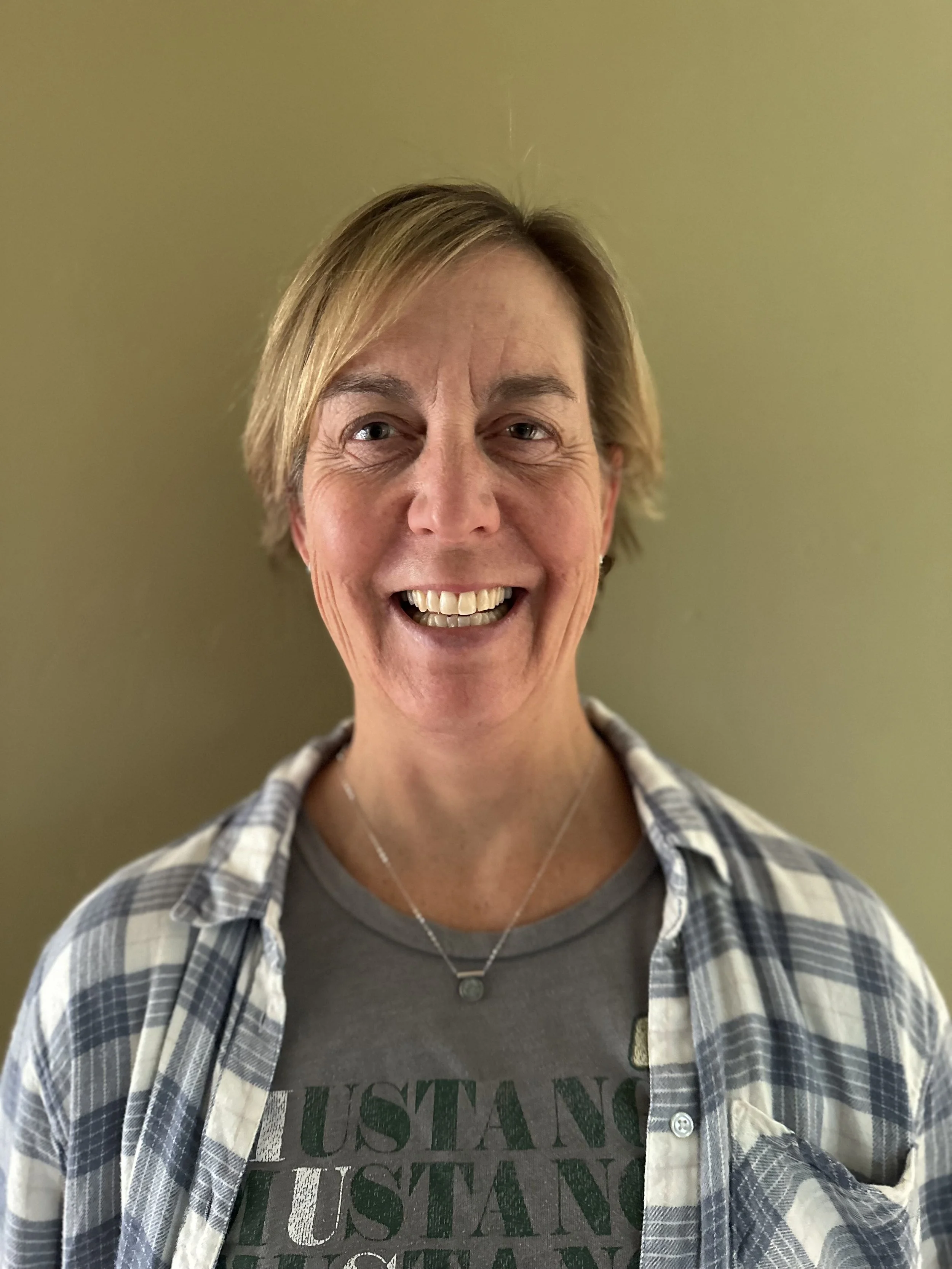 Smiling woman with short blonde hair wearing a gray t-shirt and a plaid shirt, standing against a plain wall.