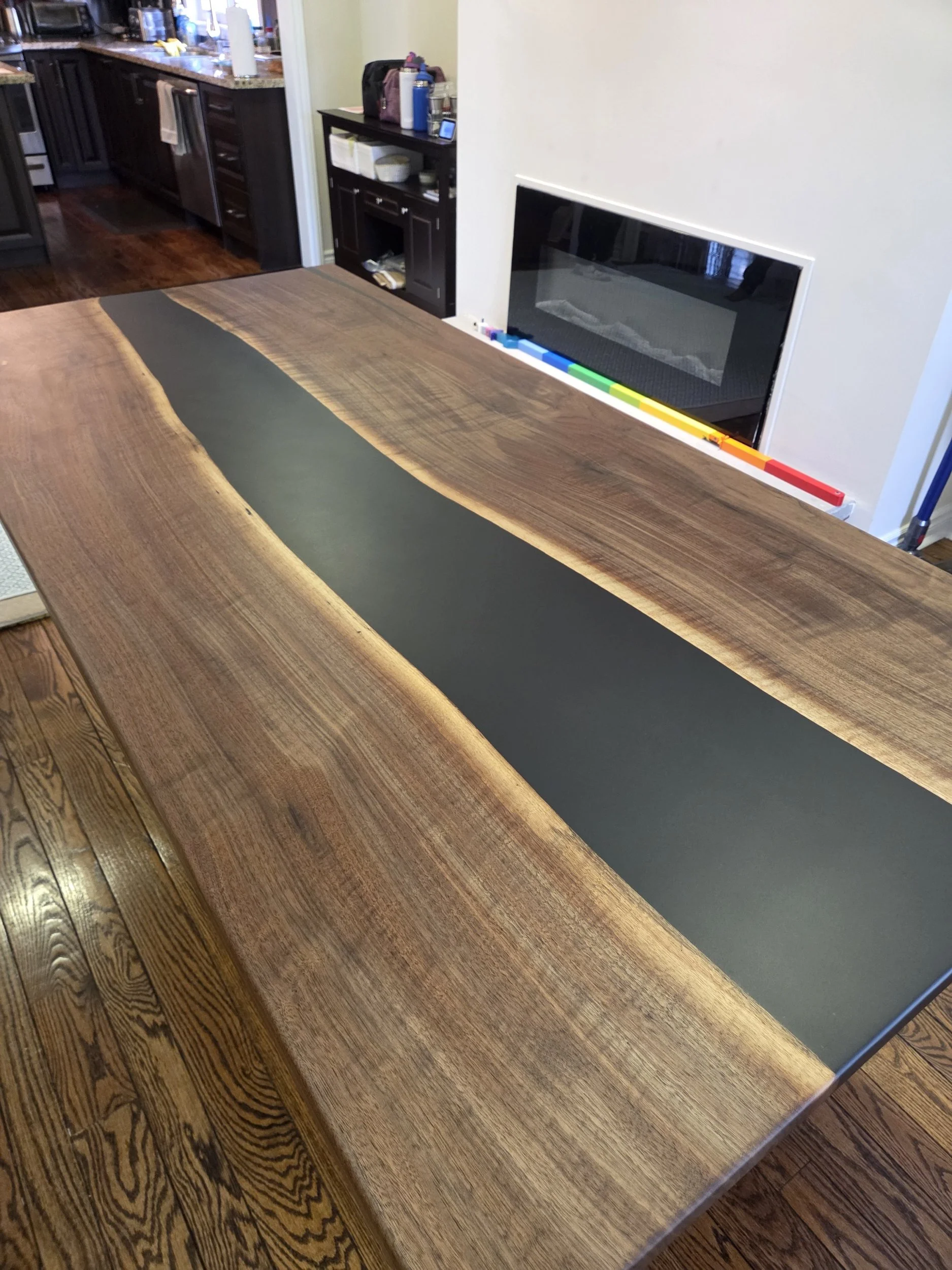 A wooden dining table with a black epoxy, situated in a kitchen with dark wood cabinets and a granite countertop.