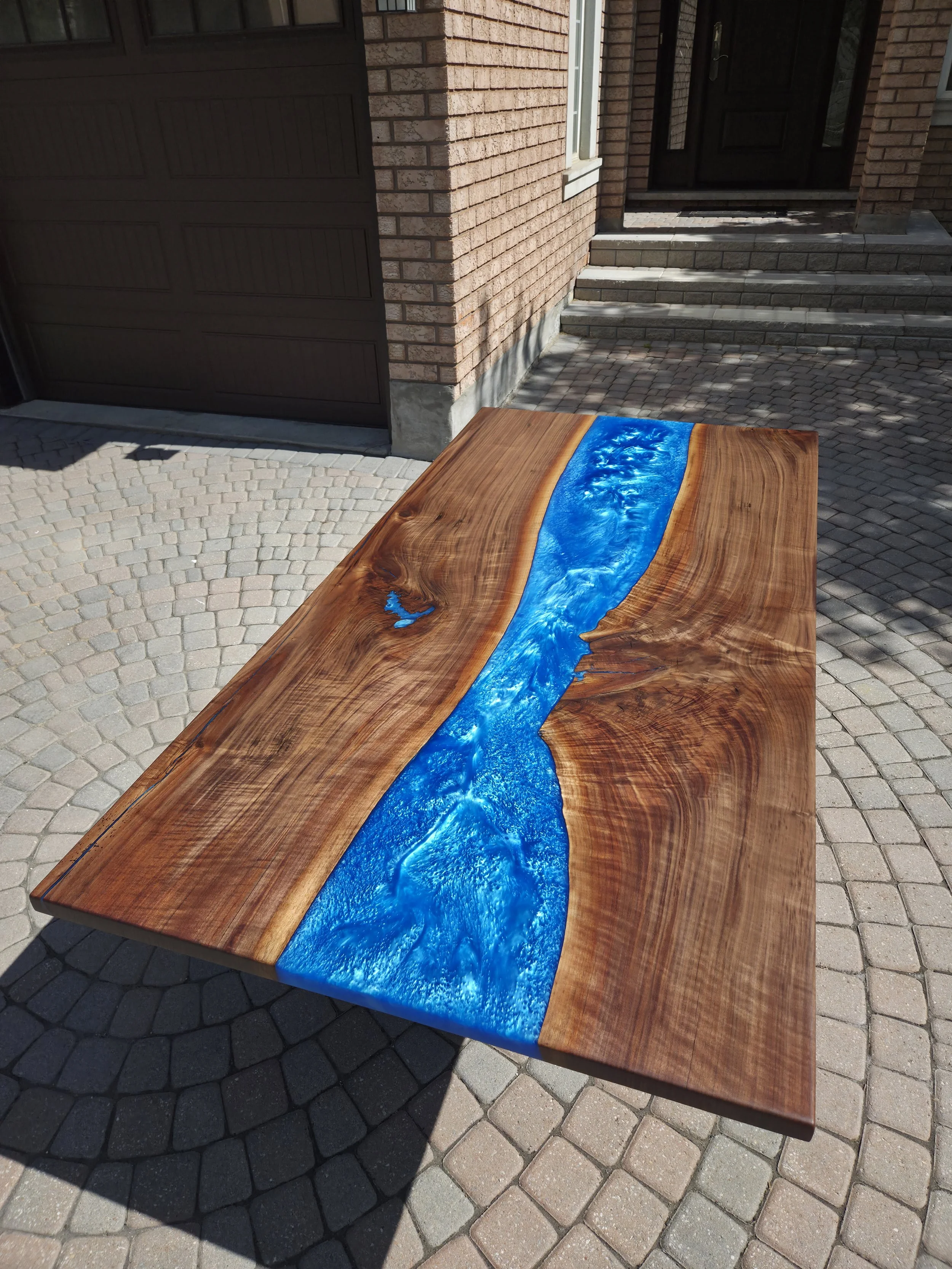 An epoxy table with a glossy, blue resin river design running through the middle of it, placed outdoors on a paved stone patio near a brick house with stairs and a garage door.