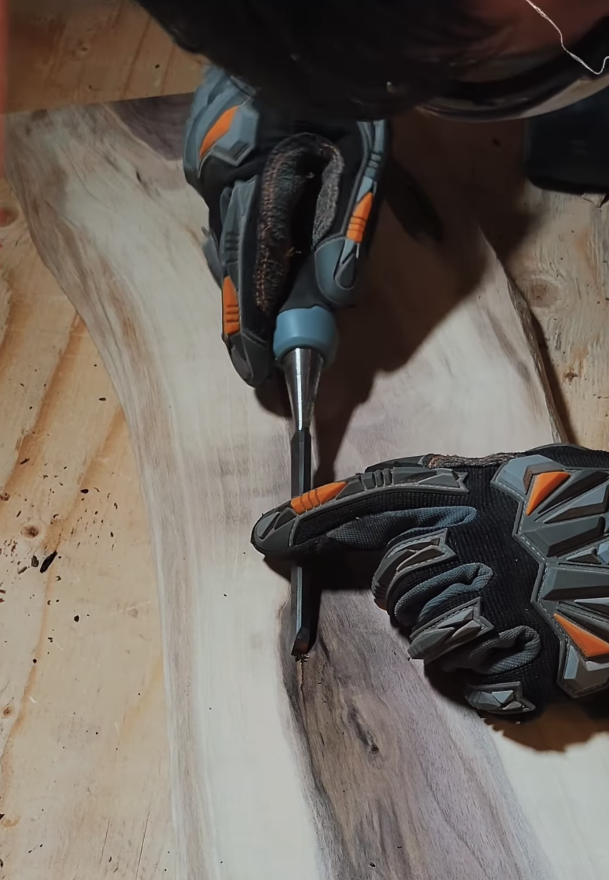 A person wearing black and orange gloves is using a chisel to work on a piece of wood. The person’s face is partially visible above the work surface.