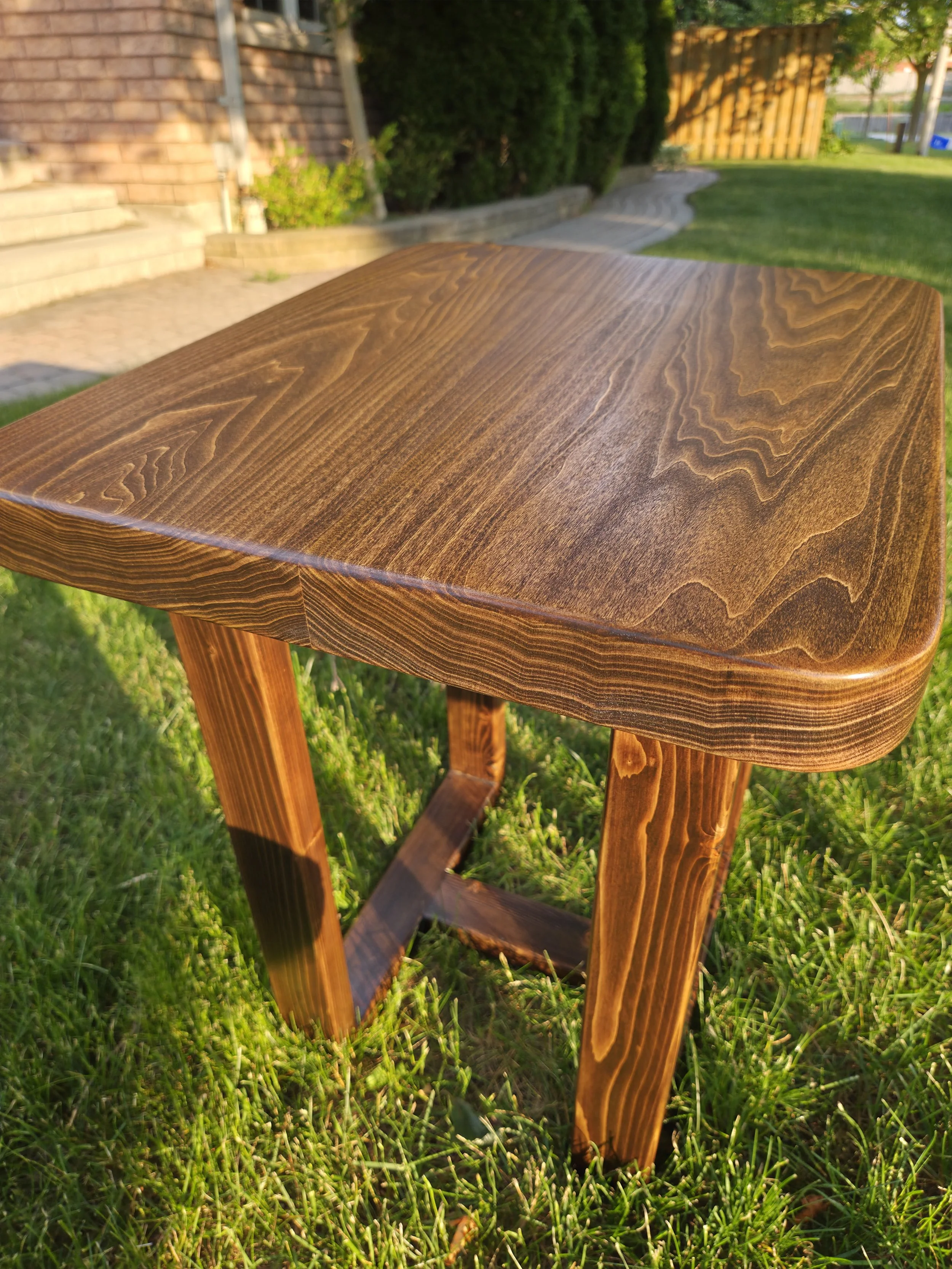 A wooden end table with a smooth, polished top and four slightly angled legs stands on green grass in a backyard, with a brick wall, bushes, a pathway, and wooden fence in the background, illuminated by sunlight.