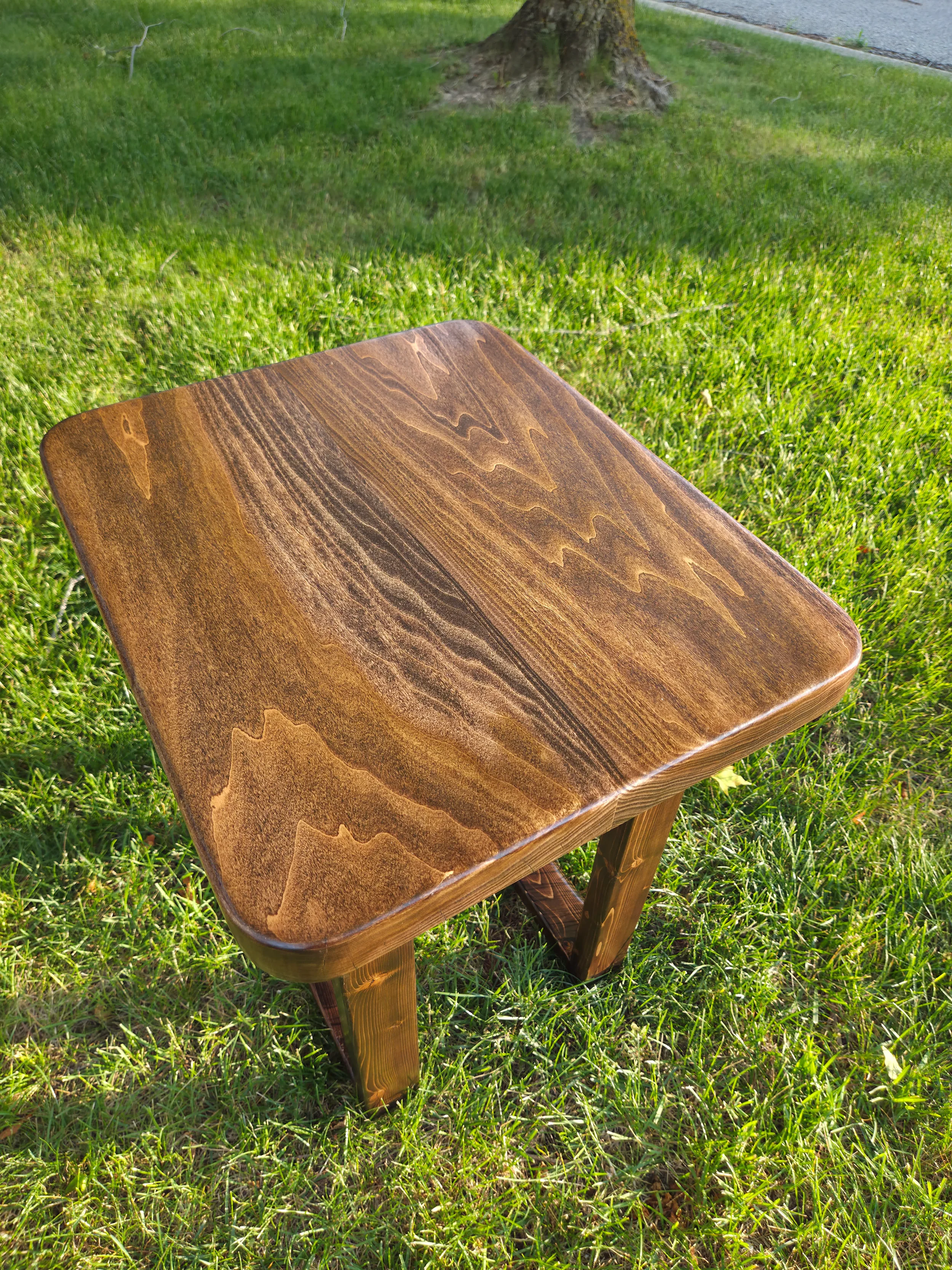 Close-up of a wooden end table outdoors on green grass, with a tree in the background.
