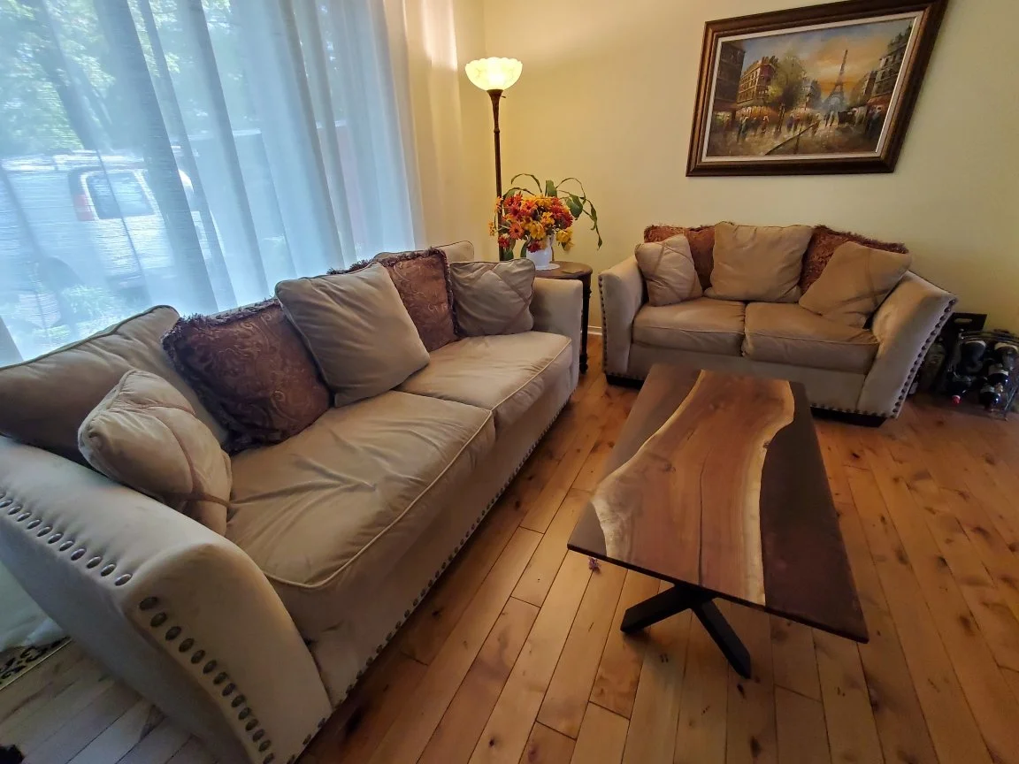 Living room with two beige sofas, an epoxy coffee table, a floor lamp, a framed painting on the wall, and a side table with flowers, next to large windows with sheer curtains.