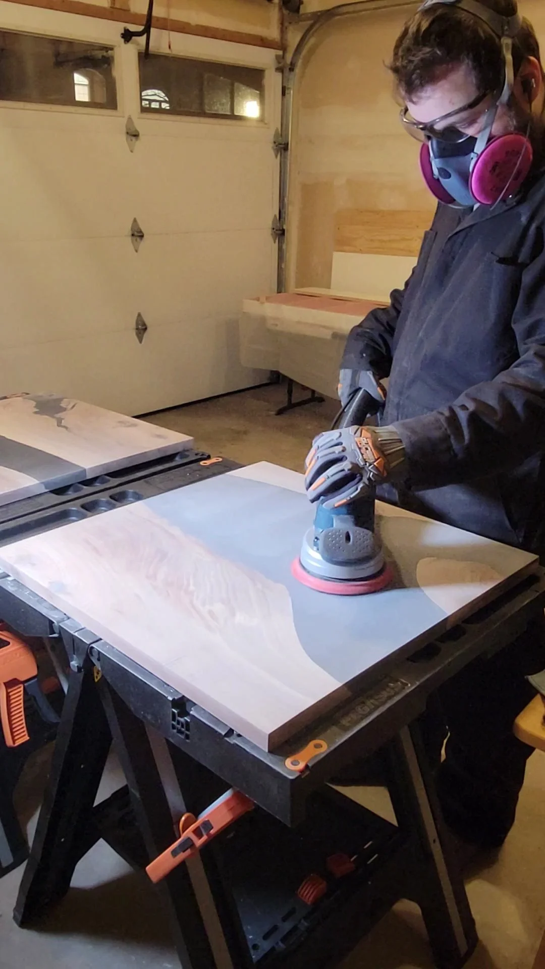 Person wearing safety goggles and a mask sanding a piece of wood with a power sander in a workshop.