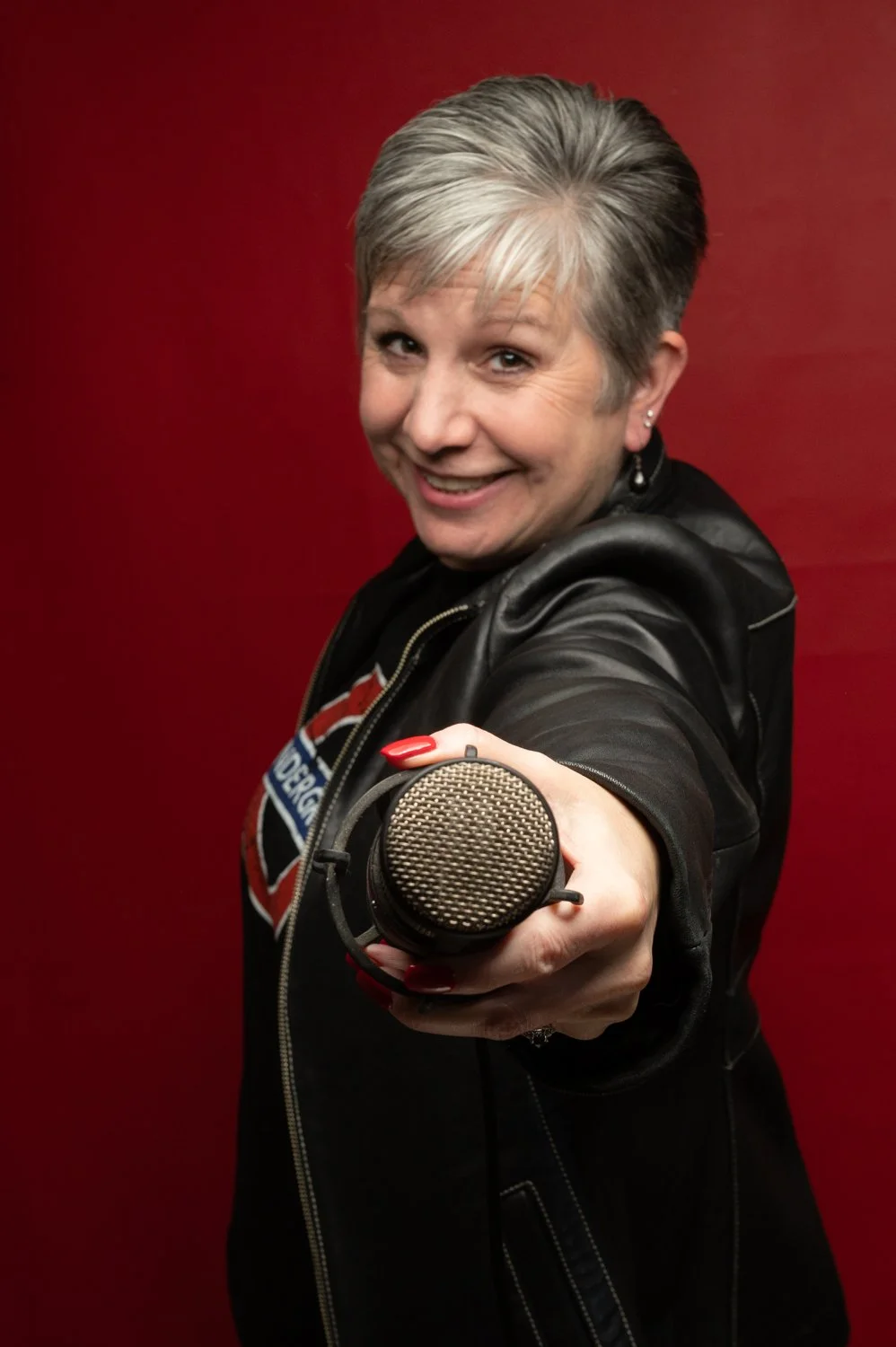 Smiling woman with short gray hair points a microphone toward the camera against a red background.