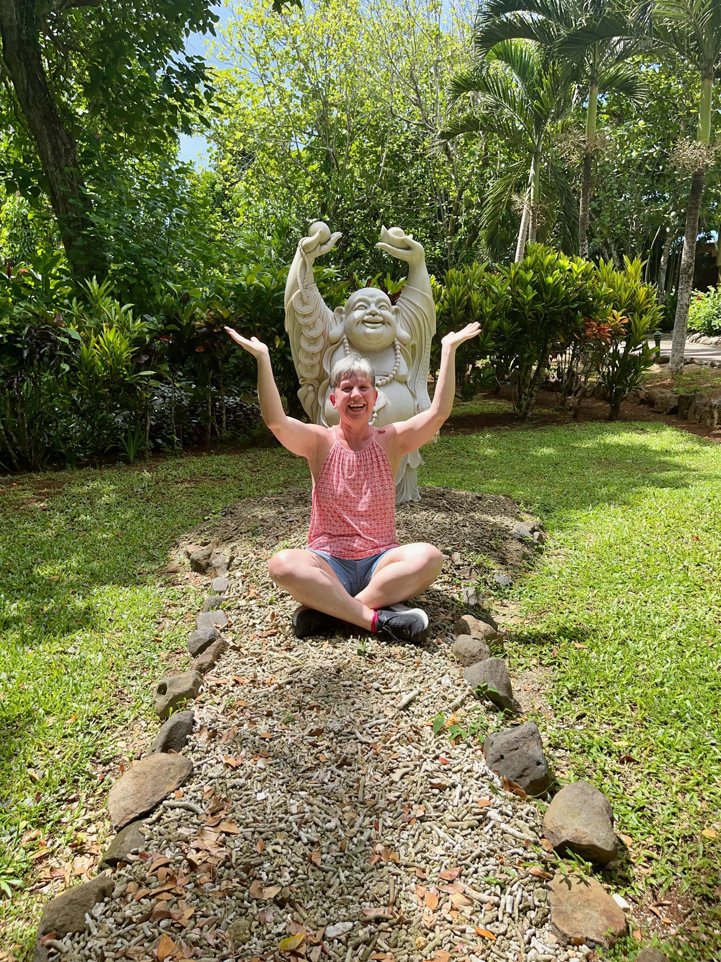 A woman sitting cross-legged on a gravel pathway with her arms raised in cheerfulness. Behind her is a large statue of a laughing, chubby figure with raised arms, set in a lush, green garden with trees and shrubs.