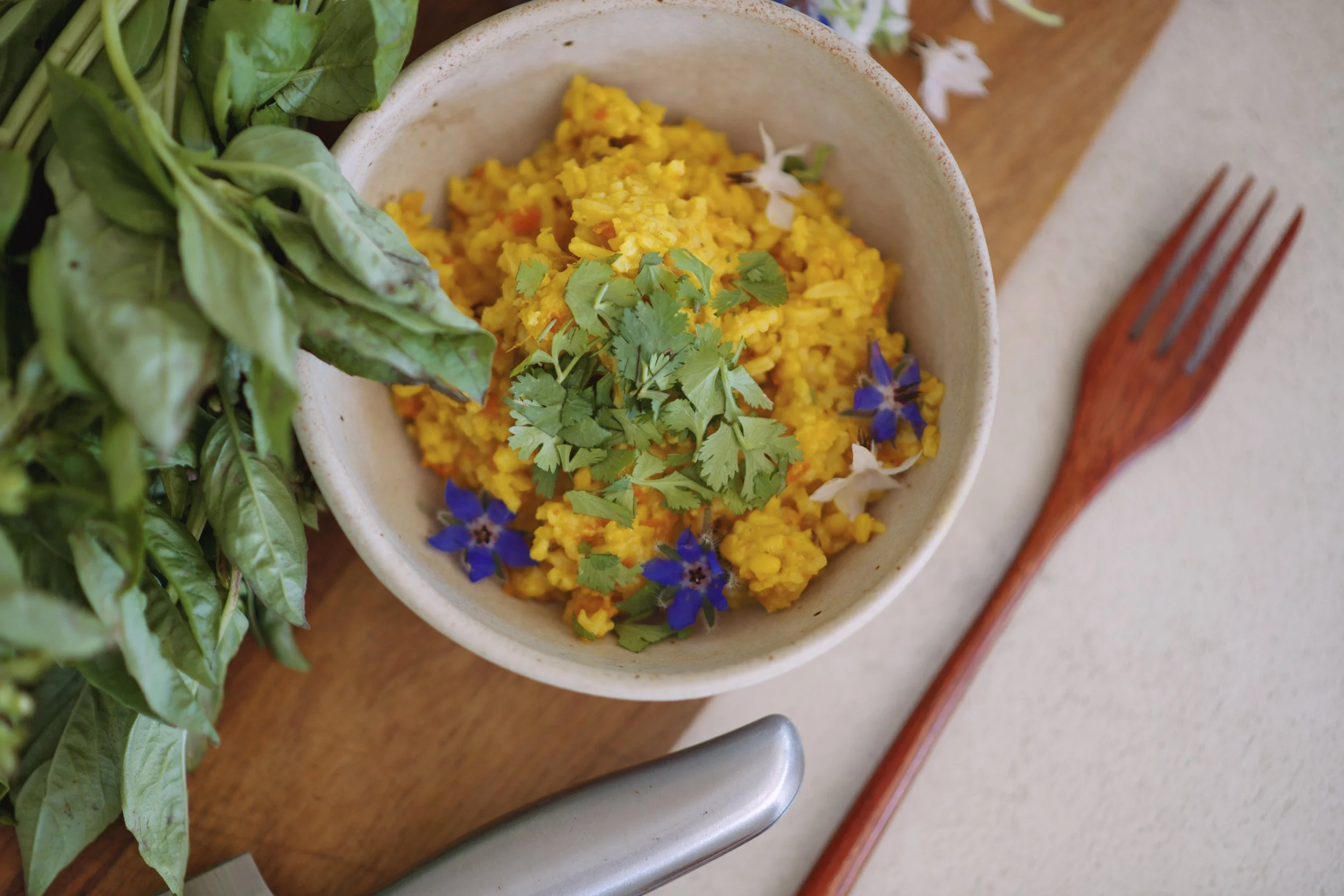 Bowl of yellow rice garnished with fresh cilantro and edible flowers, with fresh basil leaves on the side, placed on a wooden table.