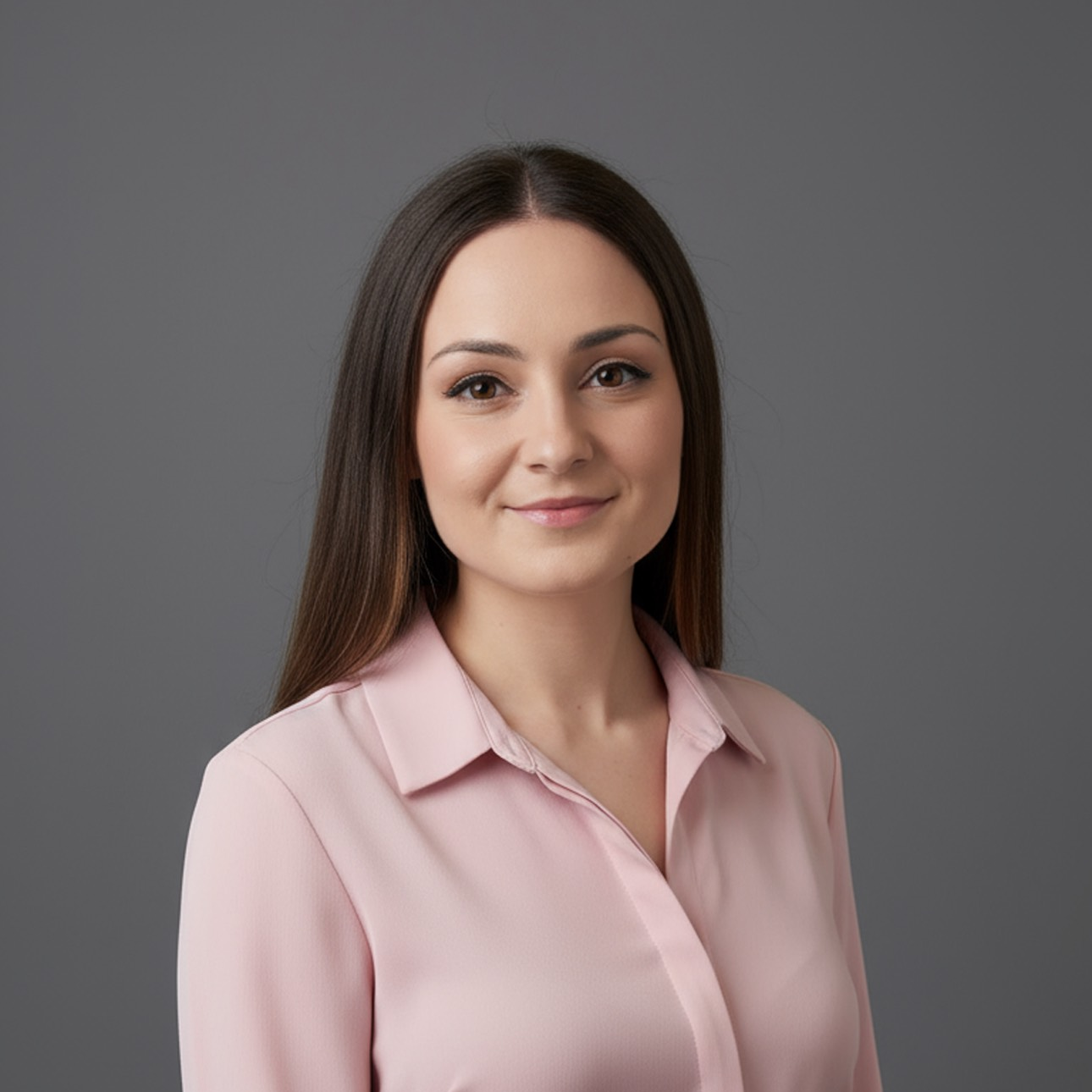 A young woman with long brown hair, wearing a light pink blouse, standing against a gray background.