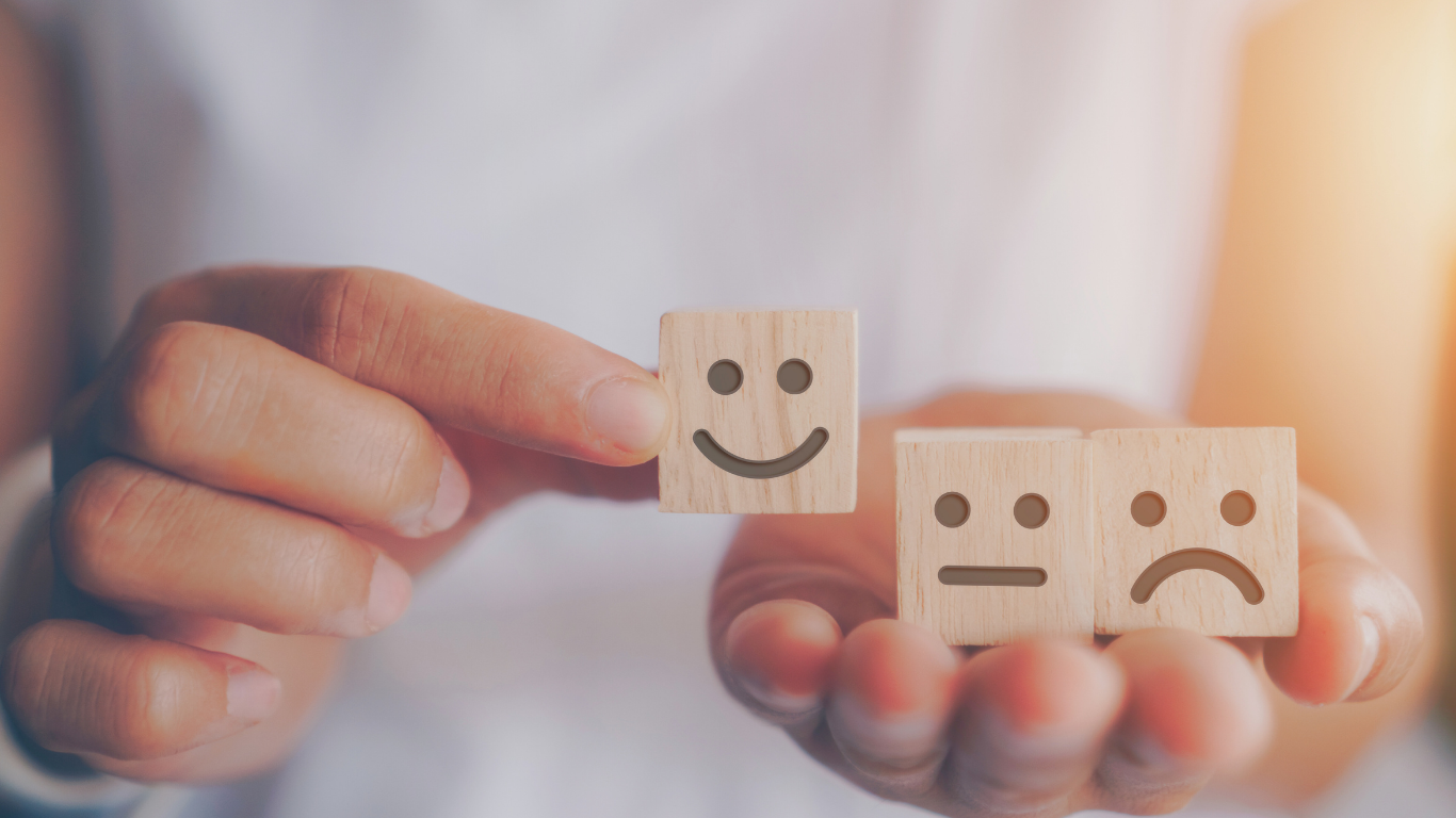 Close-up of a hand holding wooden blocks with smiley faces representing emotions: happy, neutral, and sad. Warm lighting and soft focus convey a positive tone.